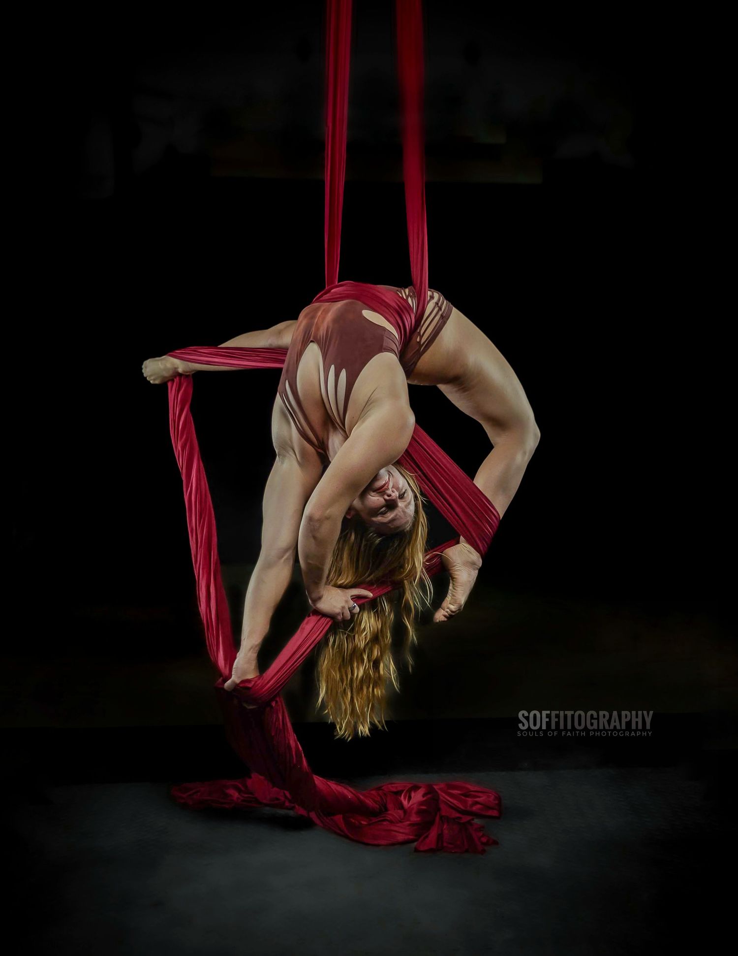 Aerial silk performer in red costume executes a dramatic backbend while suspended on red silks against dark background.