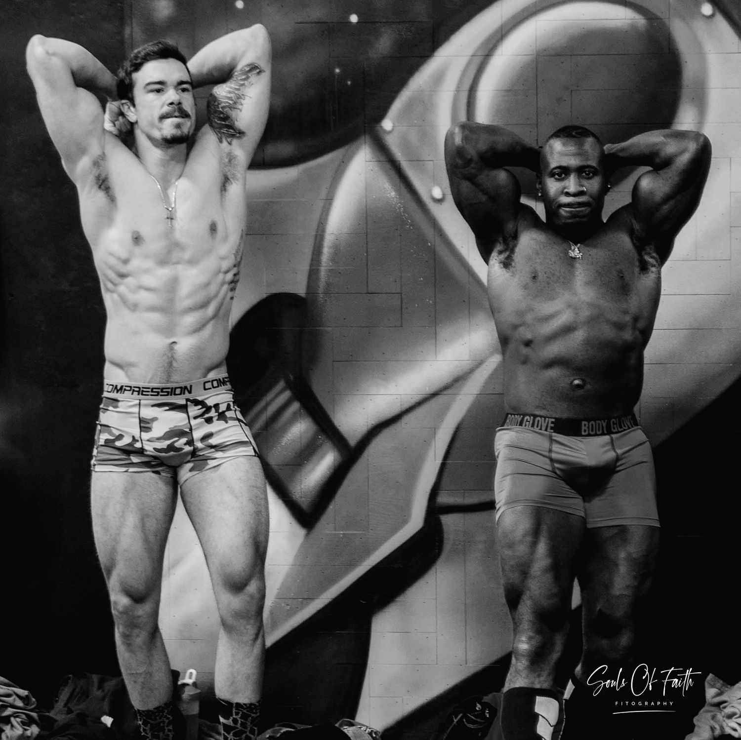 Two muscular male bodybuilders pose shirtless in black and white photo against graffiti wall backdrop.