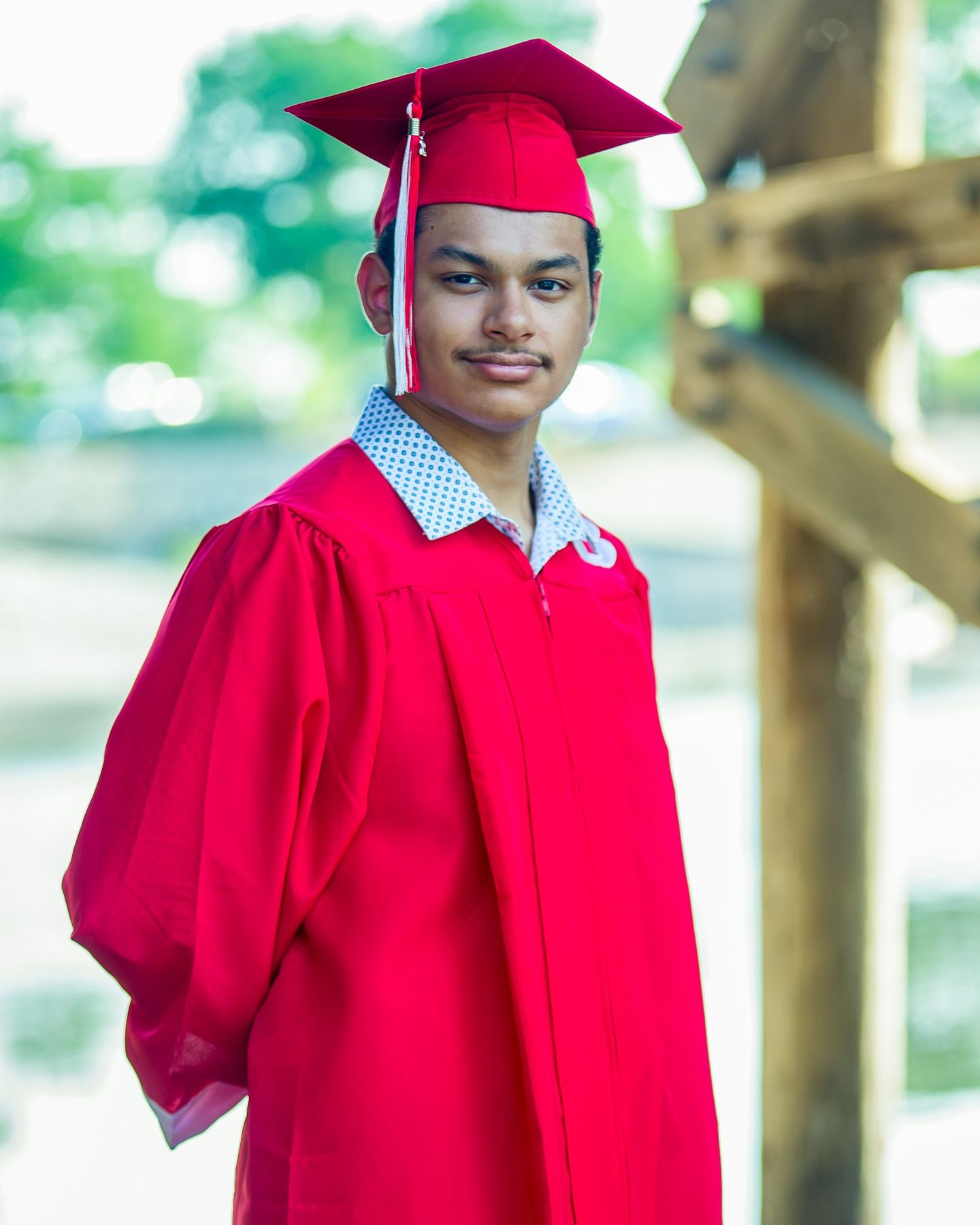 Young graduate smiling in red cap and gown outdoors on a bright sunny day.