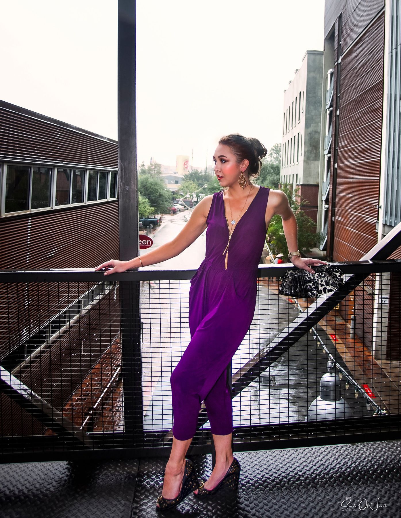 Woman in purple jumpsuit posing on urban balcony with city buildings in background.