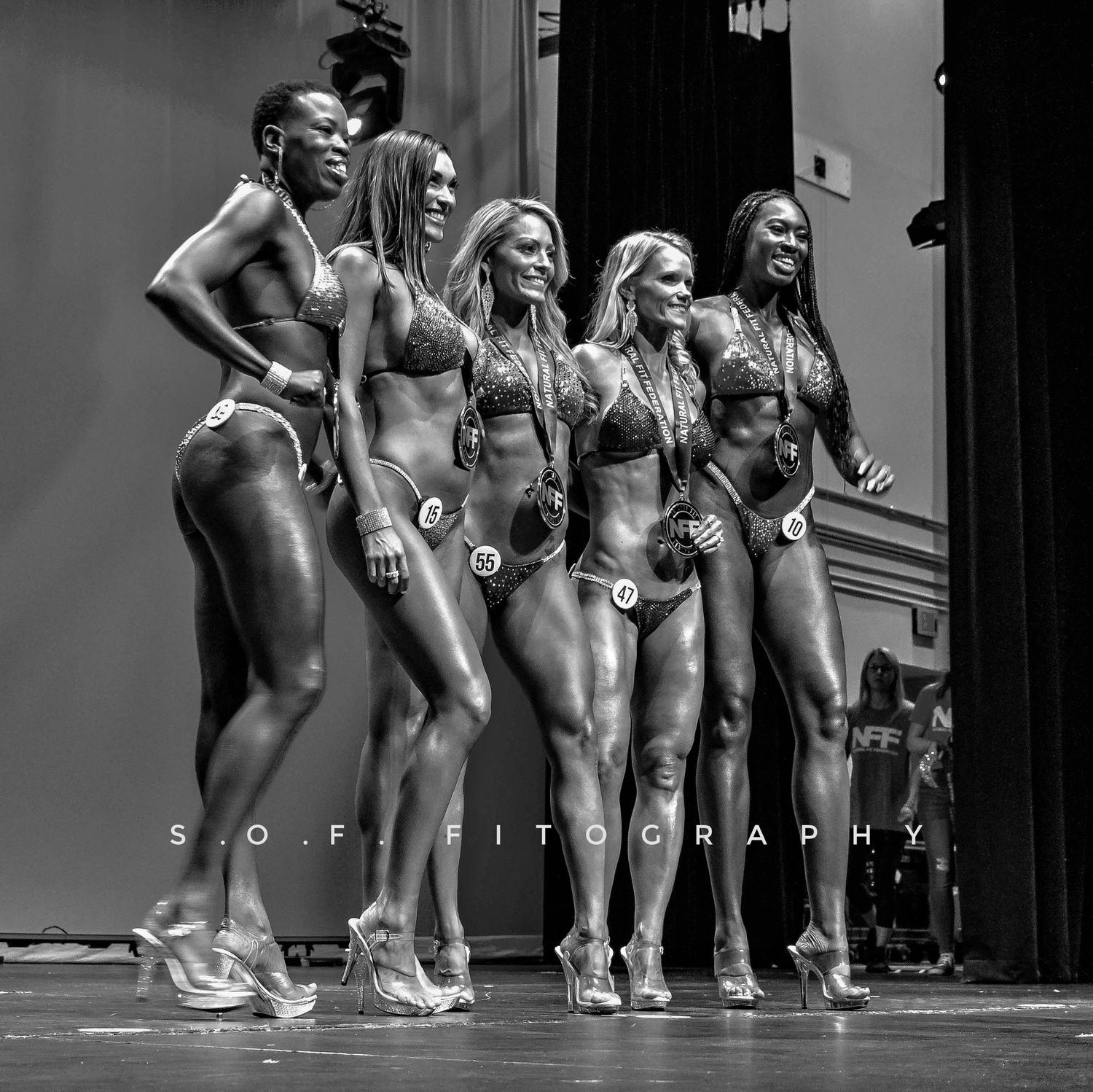 Five muscular women in bikinis pose on stage at a bodybuilding competition in this black and white photo.
