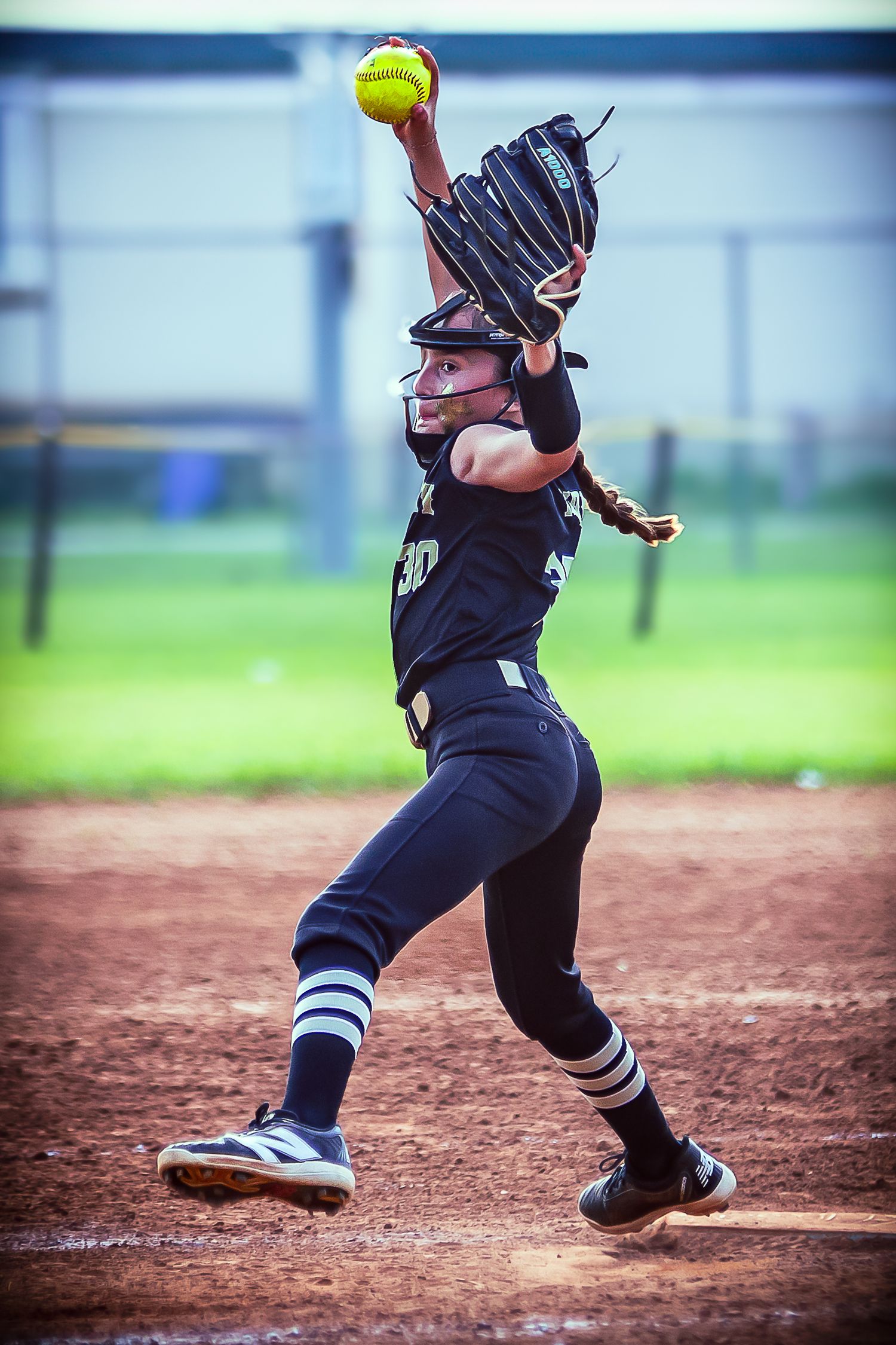 Female softball pitcher in dark uniform mid-windup, releasing yellow ball on dirt mound during game.