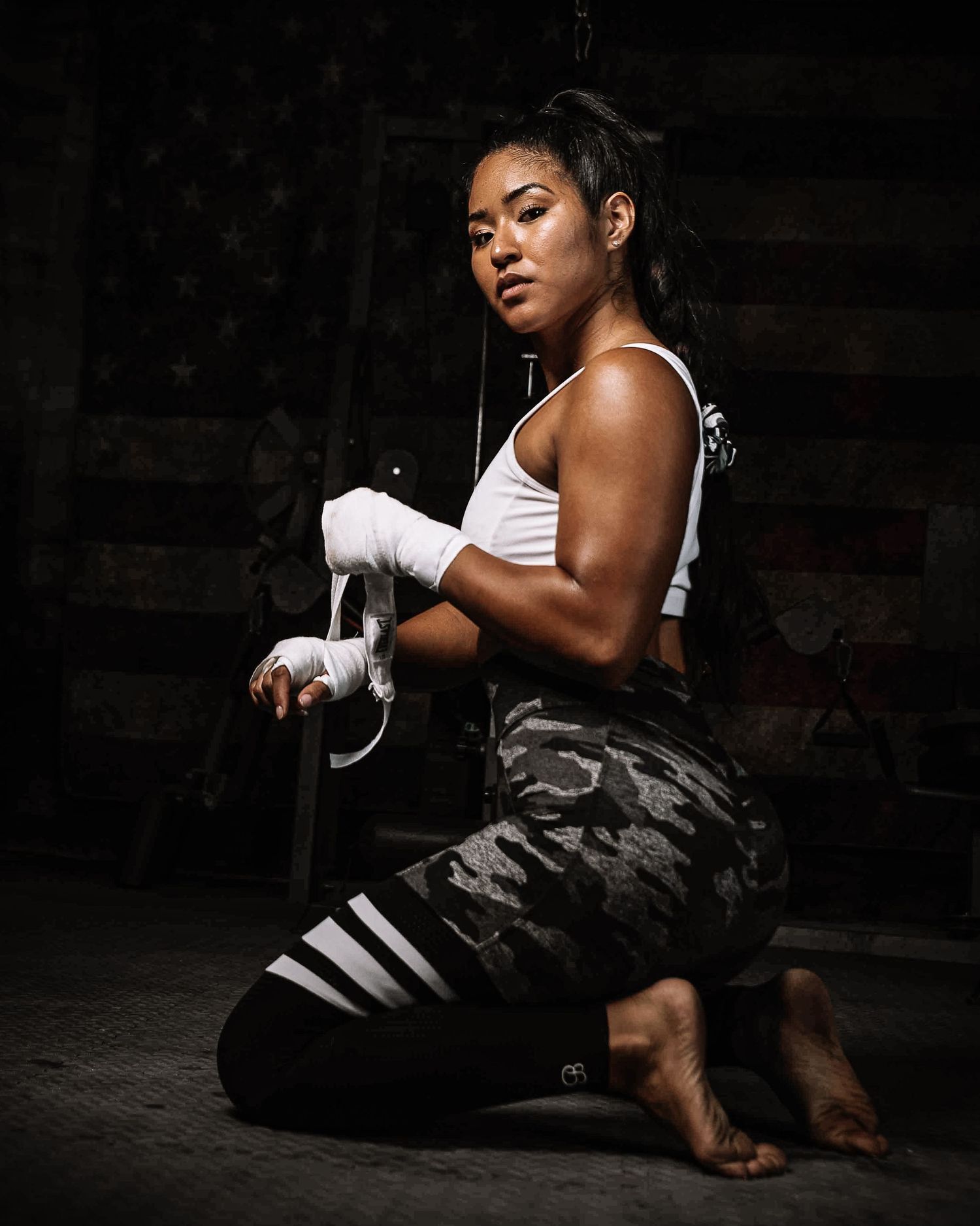 A muscular woman in camo leggings wraps her hands with boxing wraps while kneeling in a dark gym.