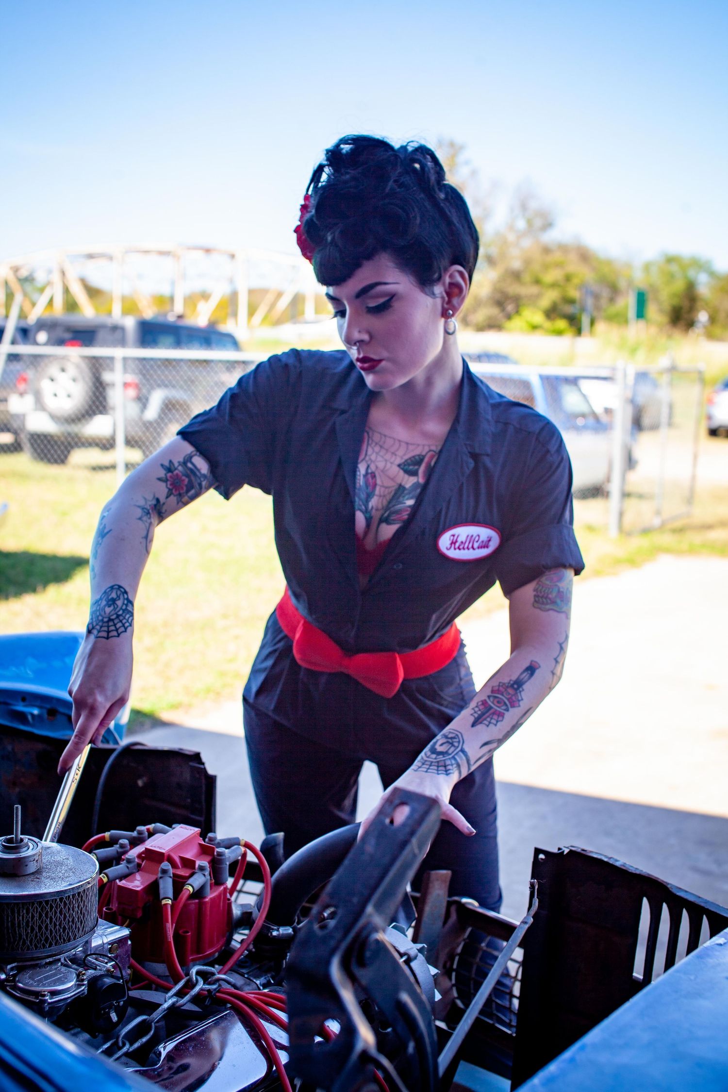 Tattooed woman in gray mechanic jumpsuit with red belt working on a car engine outdoors on a sunny day.