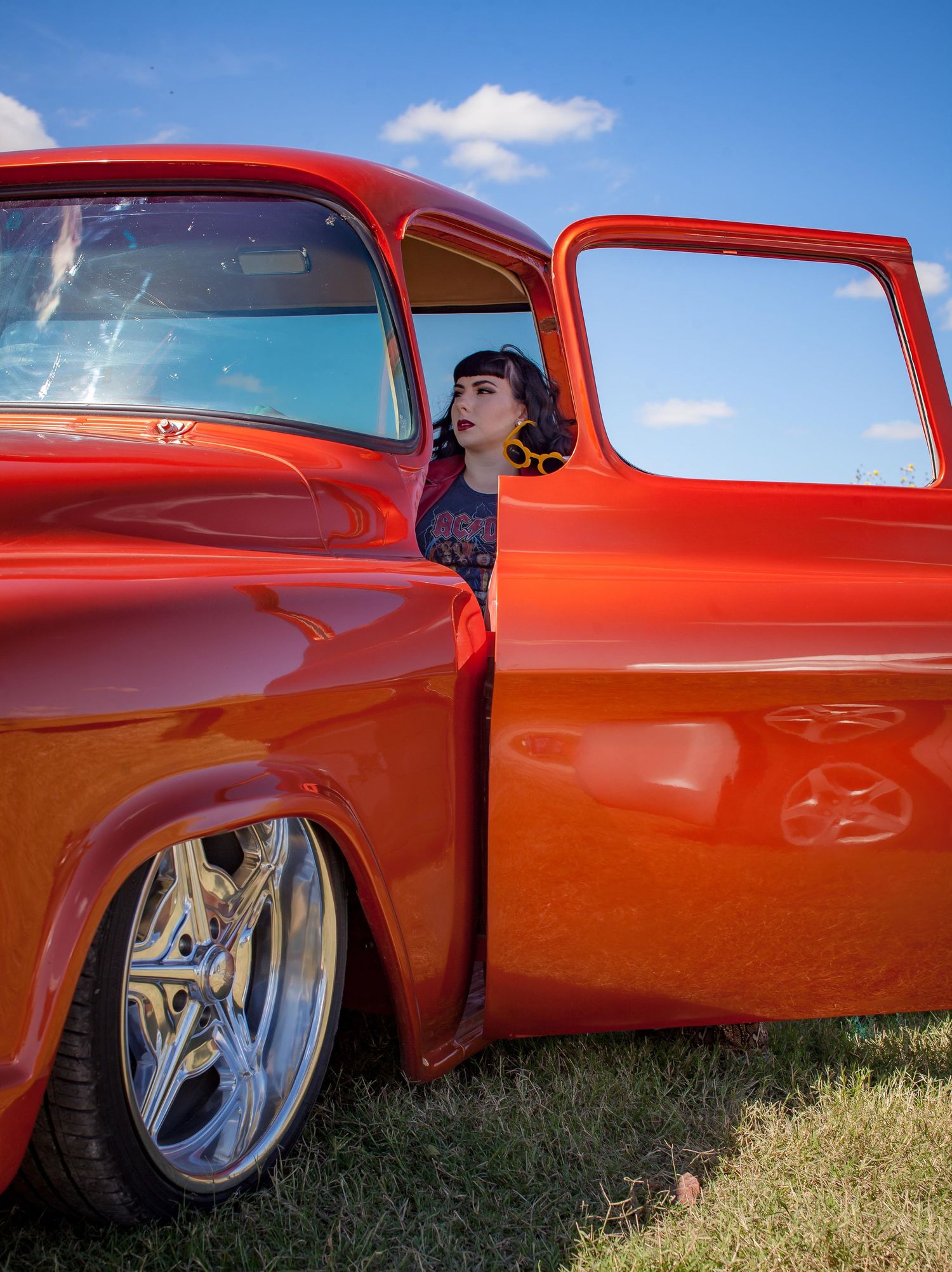 A woman peers from a stunning red custom classic truck with chrome wheels at an outdoor car show.