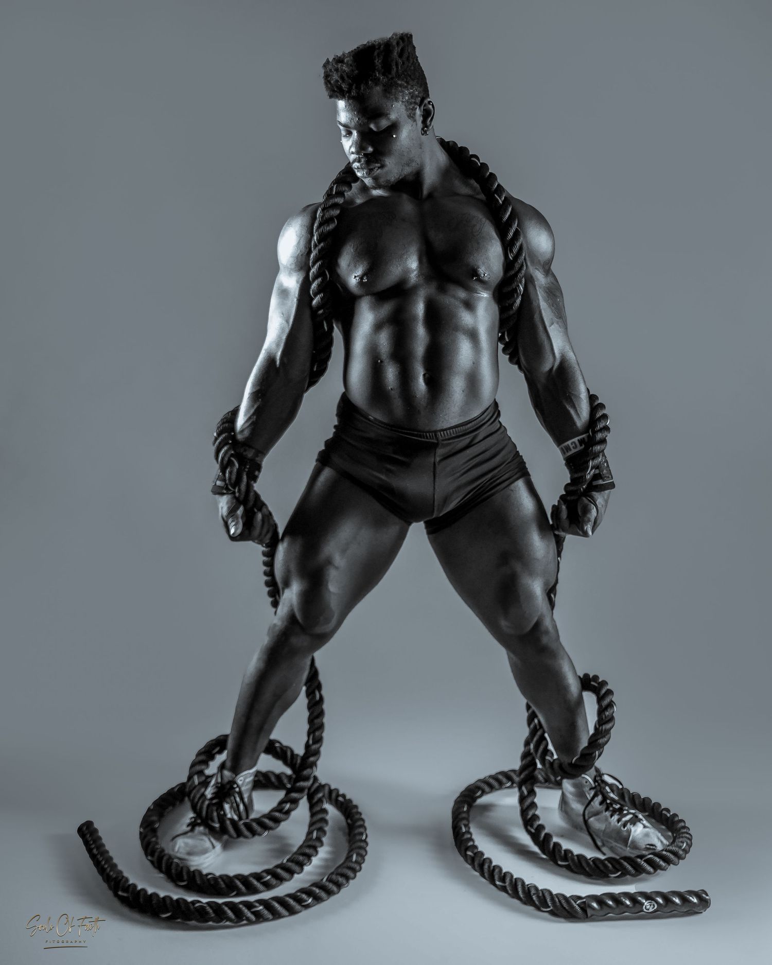 Muscular male bodybuilder posing with battle ropes in dramatic black and white studio photography.