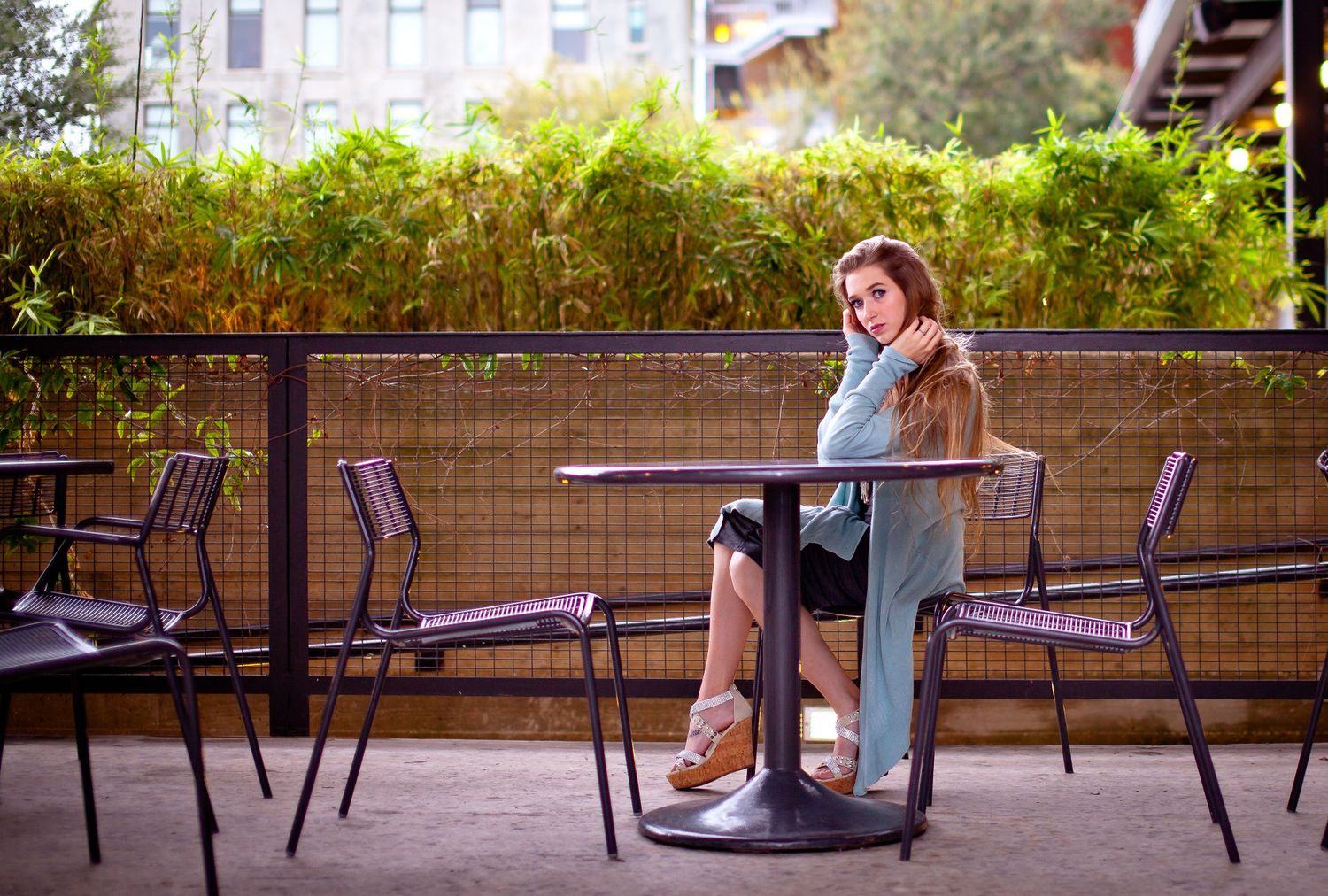 Young woman sitting alone at outdoor cafe table surrounded by lush greenery and purple chairs. Pearl lifestyle fashion