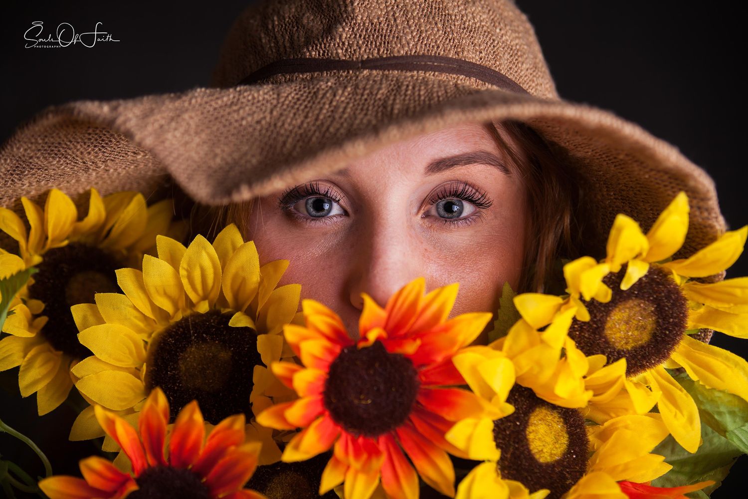 Woman with blue eyes peering over vibrant sunflowers while wearing a rustic burlap hat in a studio portrait.