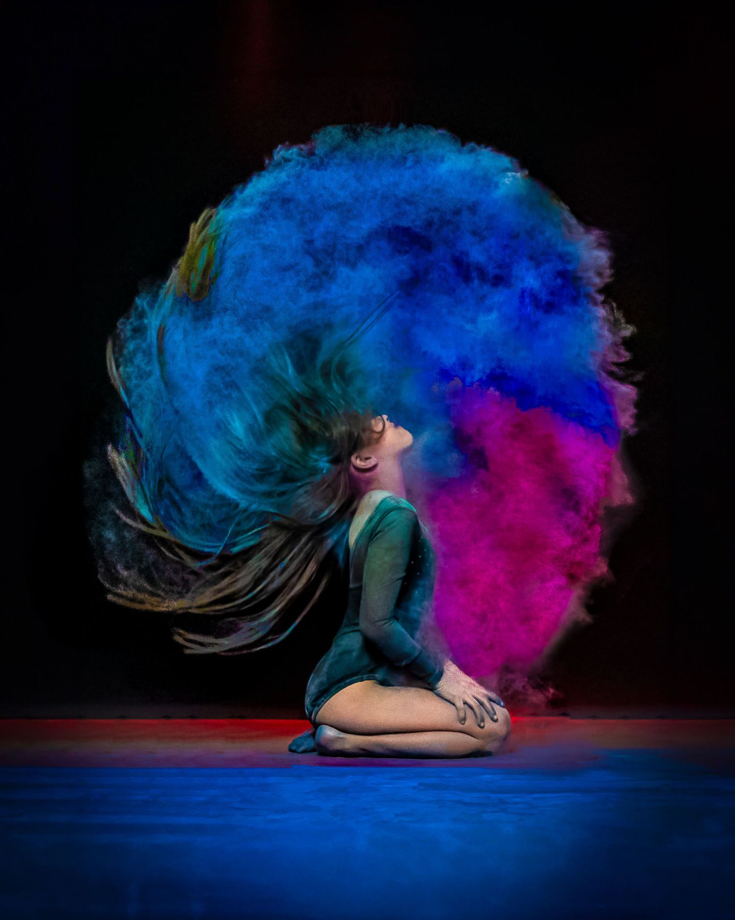 Dancer kneeling on stage with hair flipping through vibrant blue and pink powder cloud on dark background.