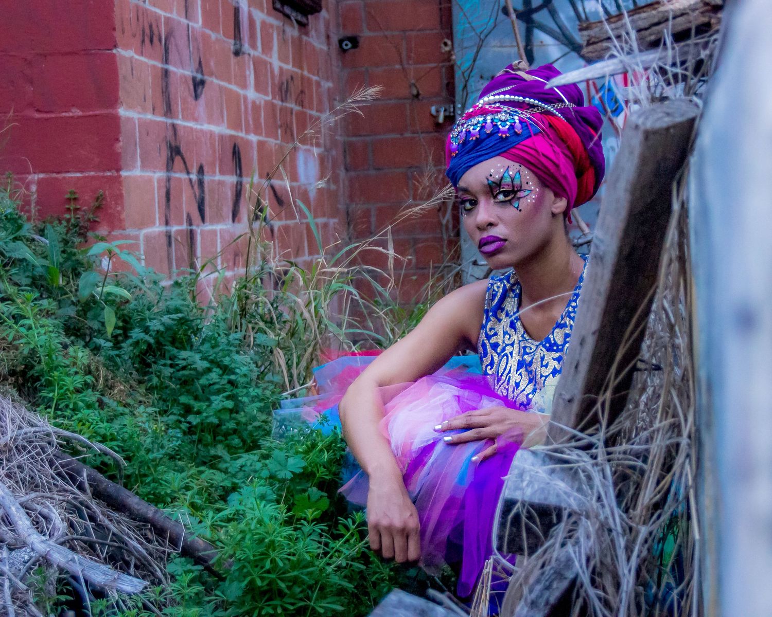 Woman in colorful turban and fantasy makeup poses in urban alleyway wearing vibrant purple tutu dress.