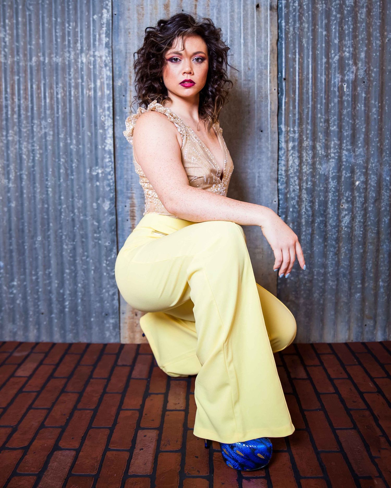 Woman in yellow wide-leg pants and beige top poses confidently on brick floor against corrugated metal wall.