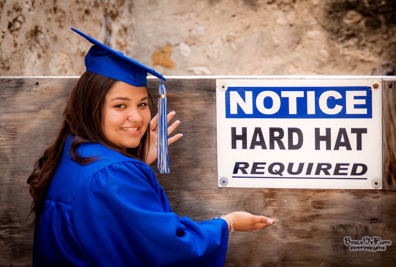 Smiling female graduate in blue cap and gown posing next to a Notice Hard Hat Required sign.