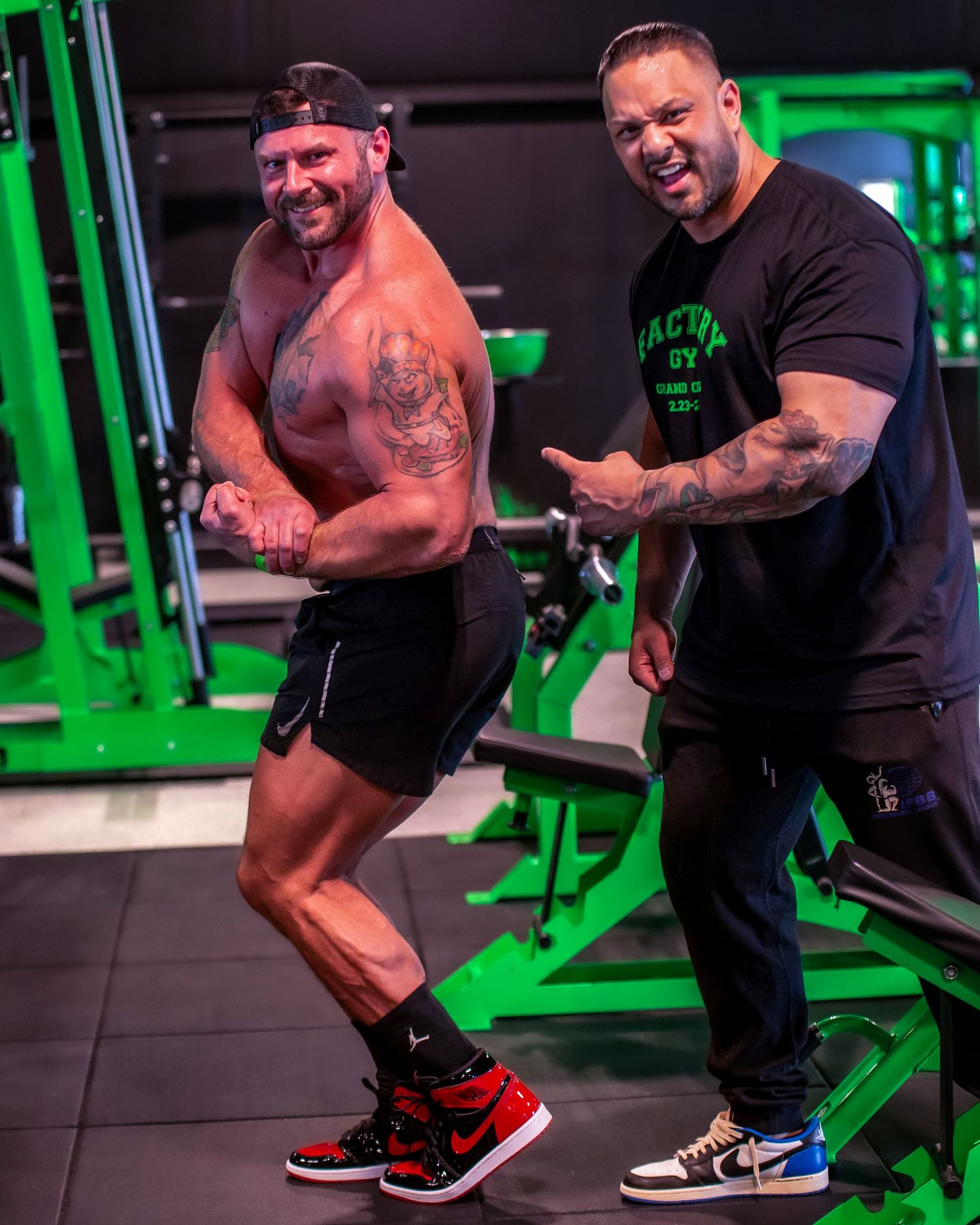 Two muscular men posing and flexing in a green-themed gym, wearing athletic wear and sneakers.