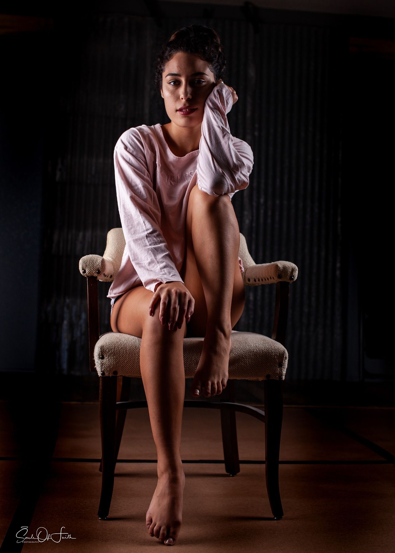 Young woman in pink shirt seated on vintage chair, barefoot, dramatic studio lighting against dark corrugated background.