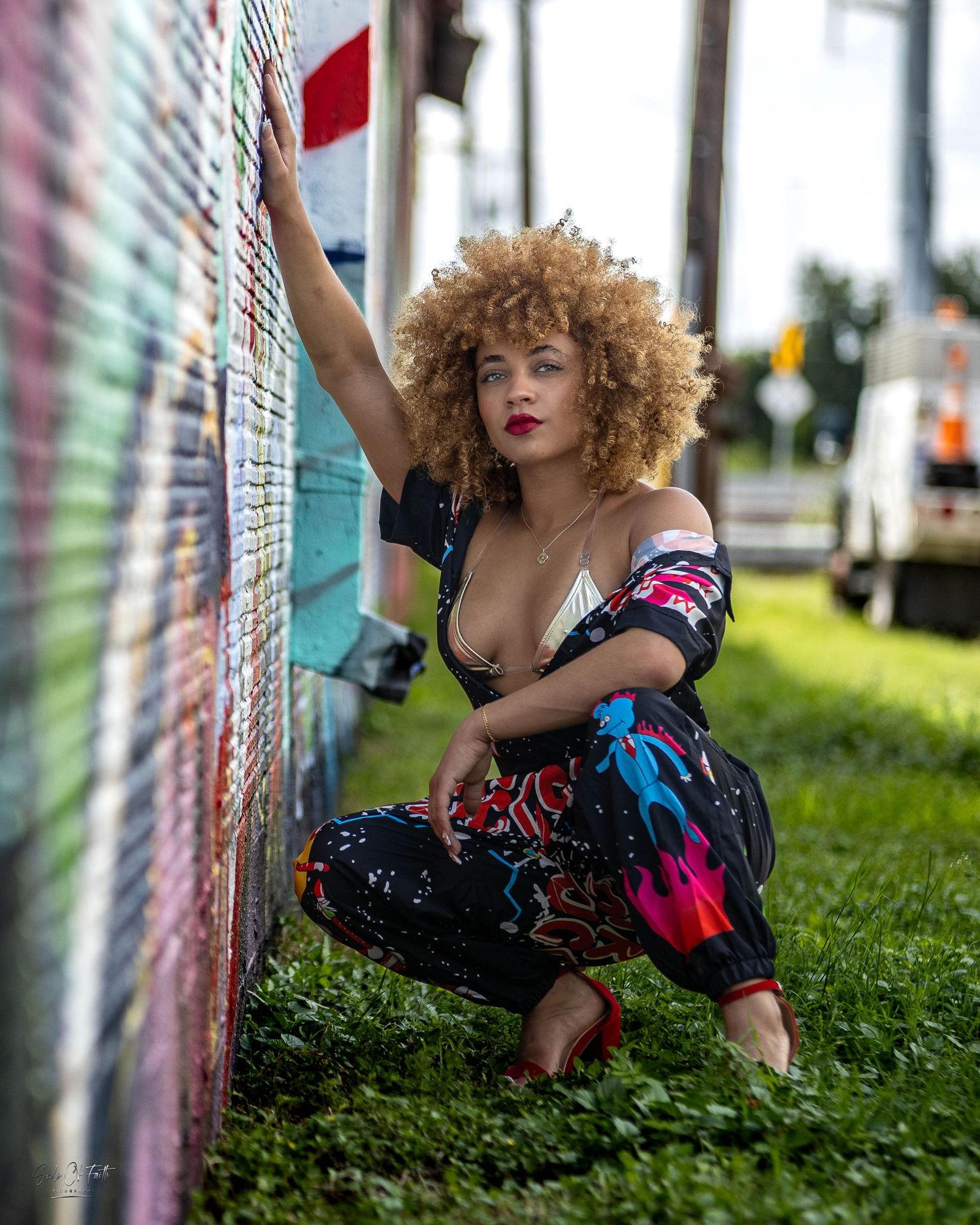 Woman with curly blonde hair in colorful outfit posing by graffiti wall on green grass.