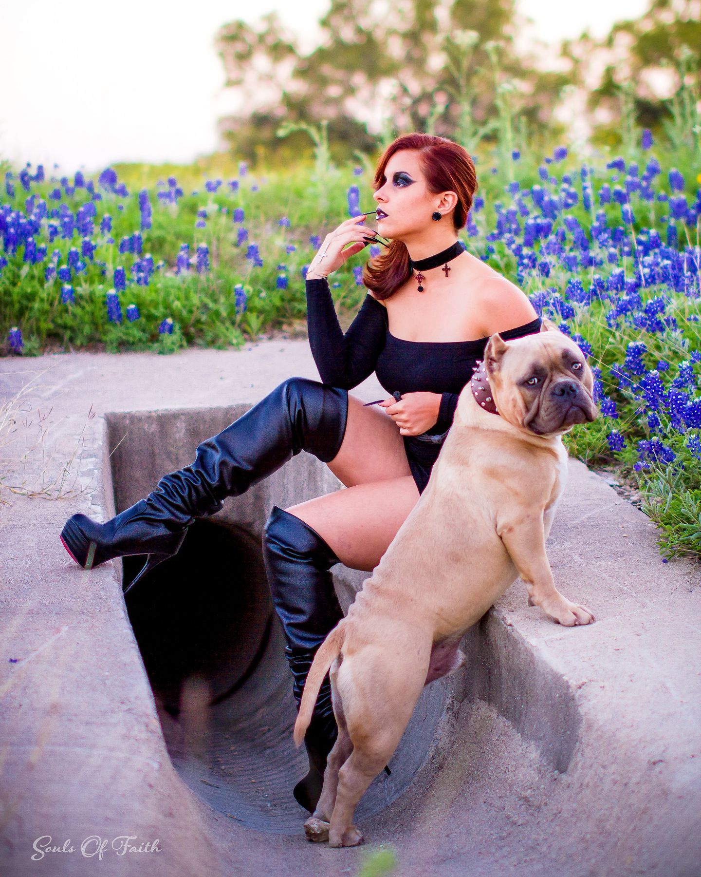 Woman in black bodysuit and thigh-high boots poses with a large tan bulldog among blue wildflowers.