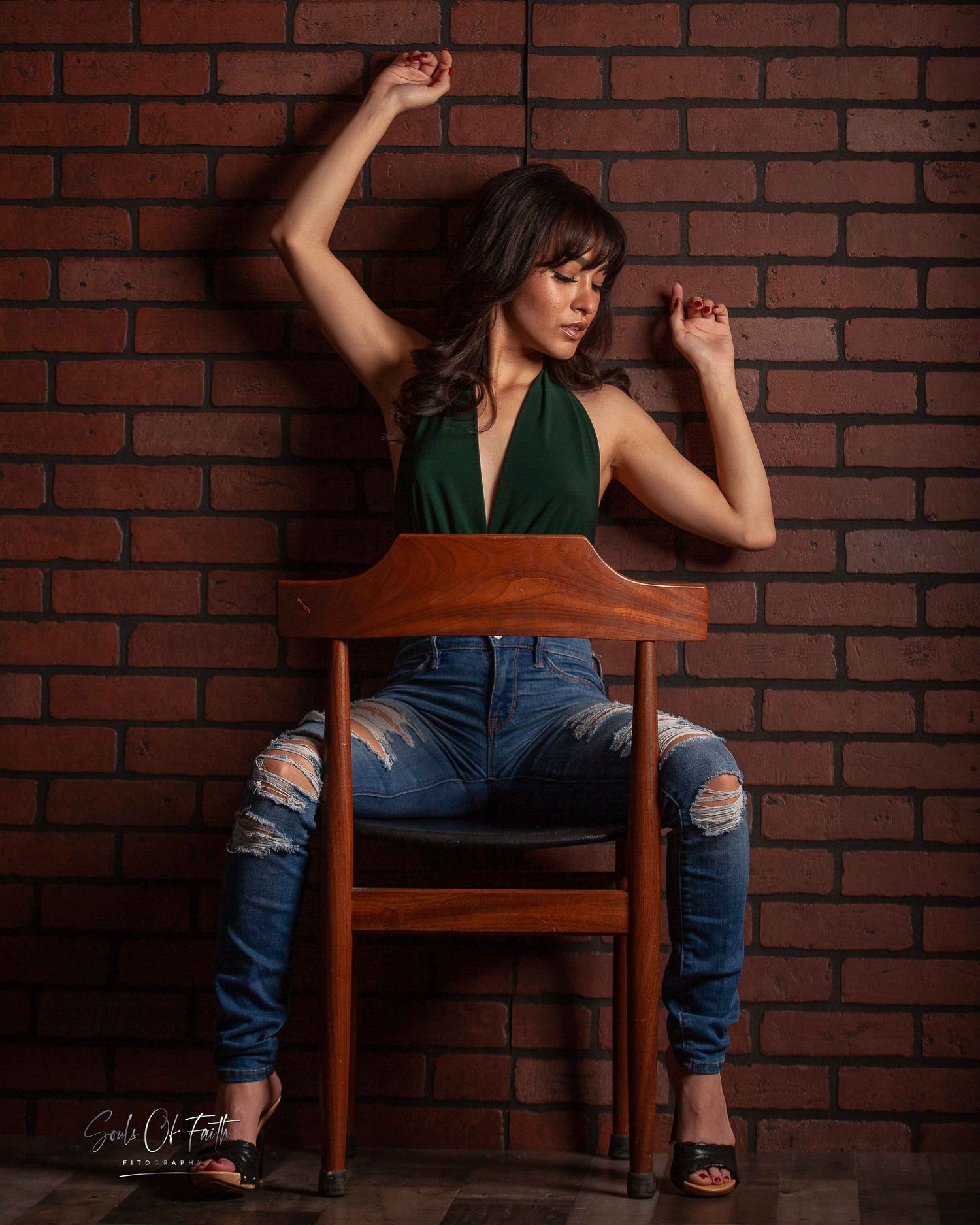 Woman in green halter top and ripped jeans poses on wooden chair against rustic brick wall.