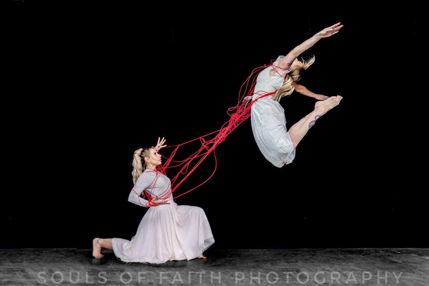 Two dancers perform an expressive contemporary dance, one leaping mid-air while connected by flowing red ribbons against a dark backdrop.
