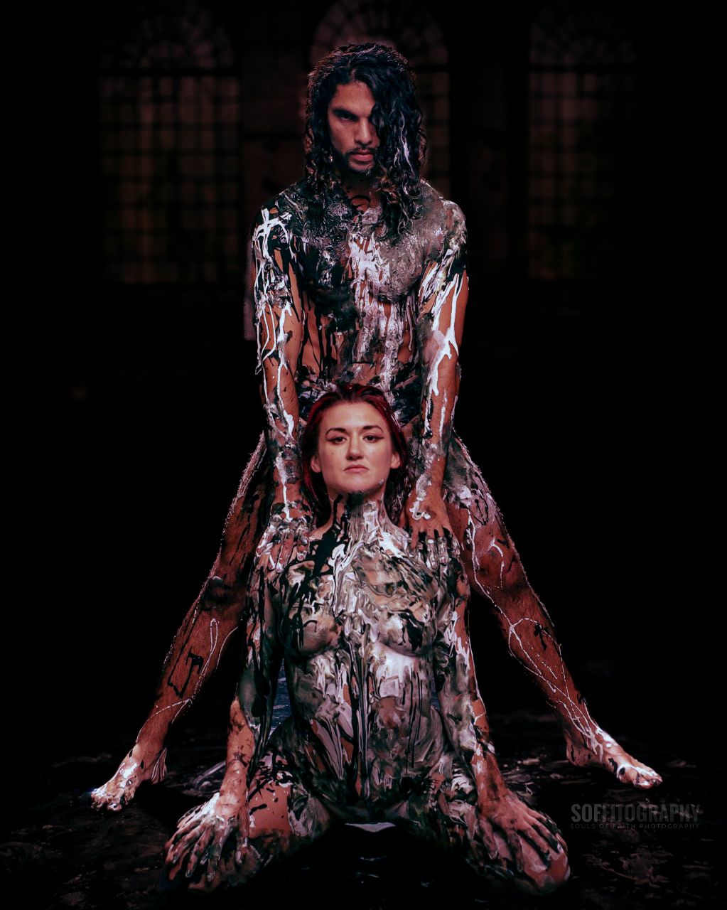 Two models covered in dripping paint pose dramatically in a dark, artistic body paint photography session.