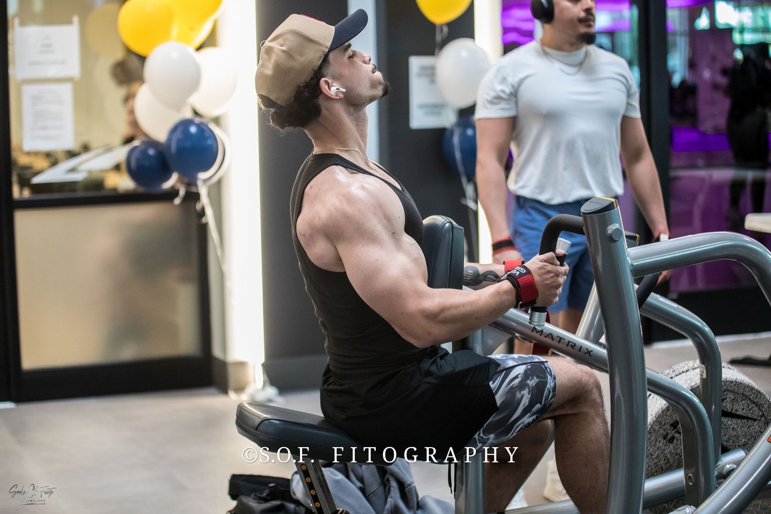 Muscular man exercising on gym machine, wearing tank top and cap, with balloons in background.