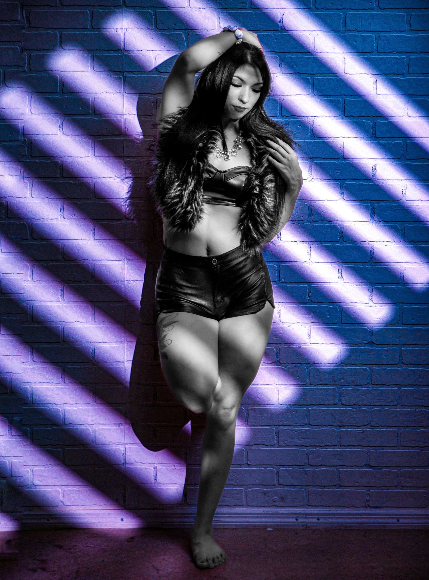 A curvy woman poses confidently in black leather shorts and crop top against a purple-lit brick wall.
