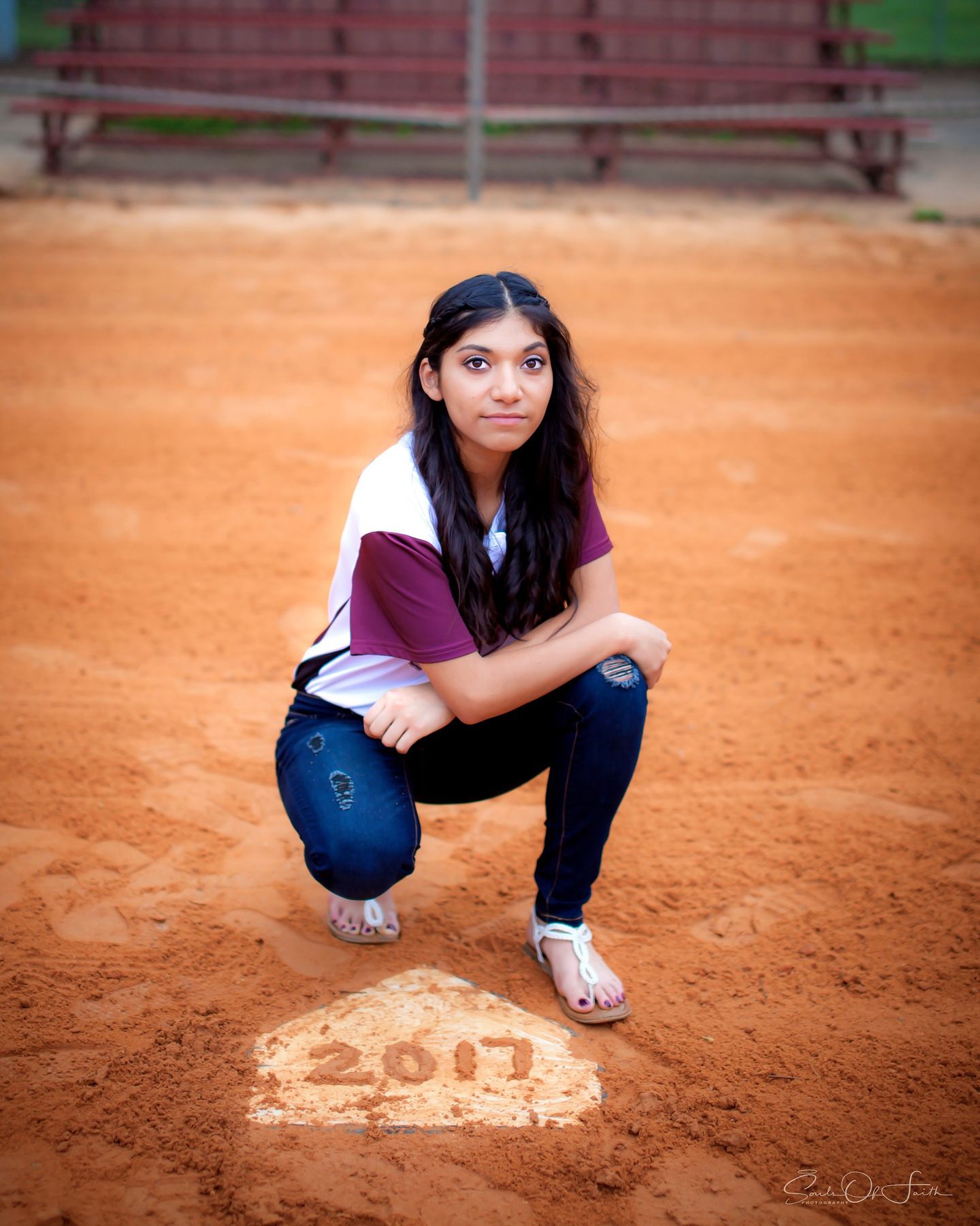 Teen girl crouching on baseball home plate marked 2017, wearing jeans and white top, on a red clay field.