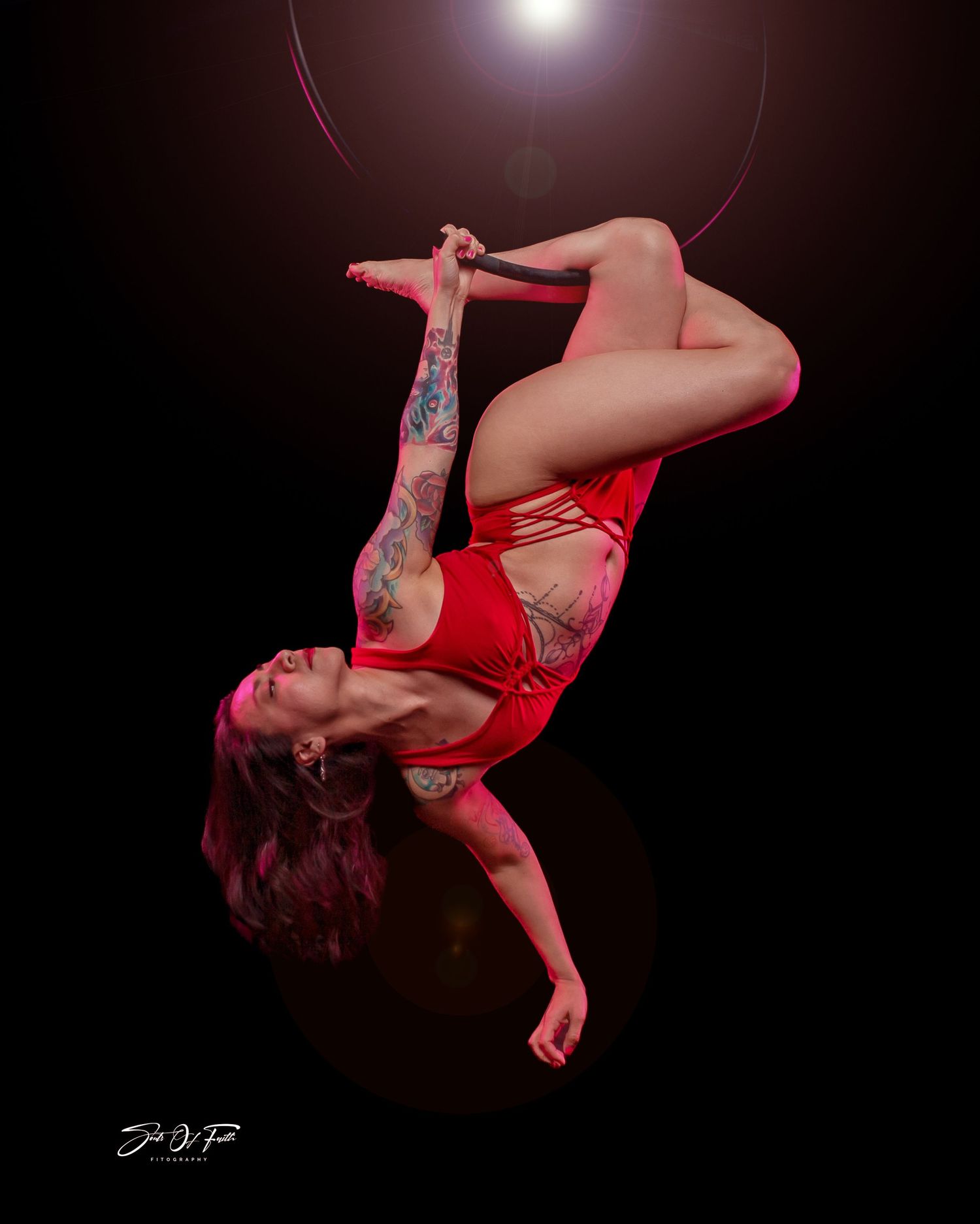 Tattooed aerial hoop performer in red bodysuit executing an inverted pose against dark dramatic background.
