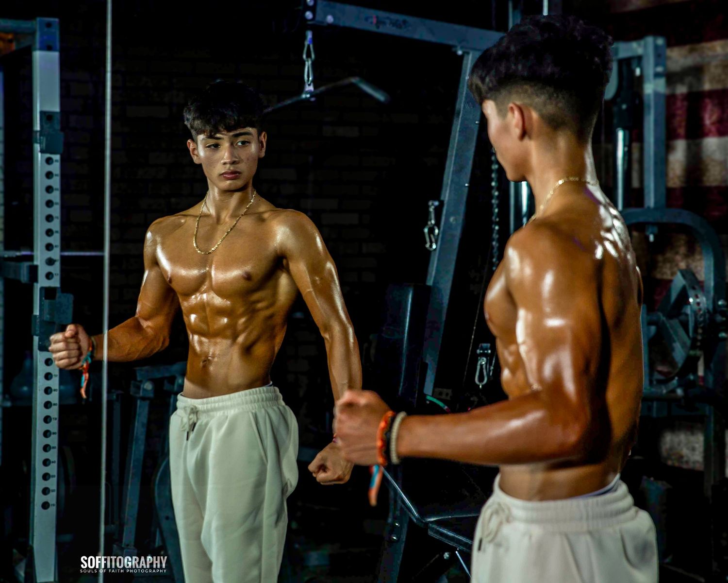 Muscular young man doing cable exercises shirtless in a gym, reflected in mirror, showing defined abs.