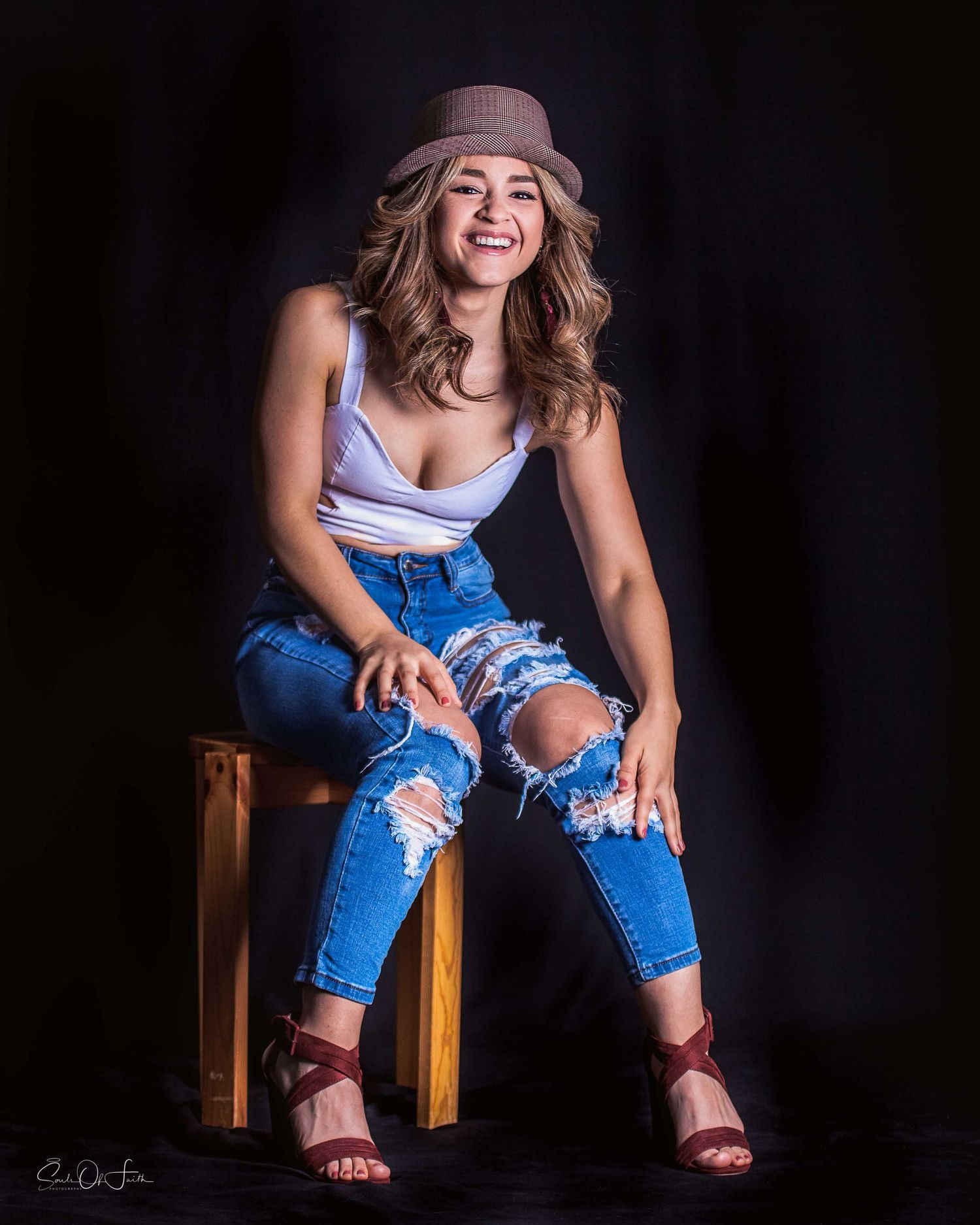 Smiling woman in ripped jeans, white top, and fedora hat sitting on a wooden stool against a dark background.