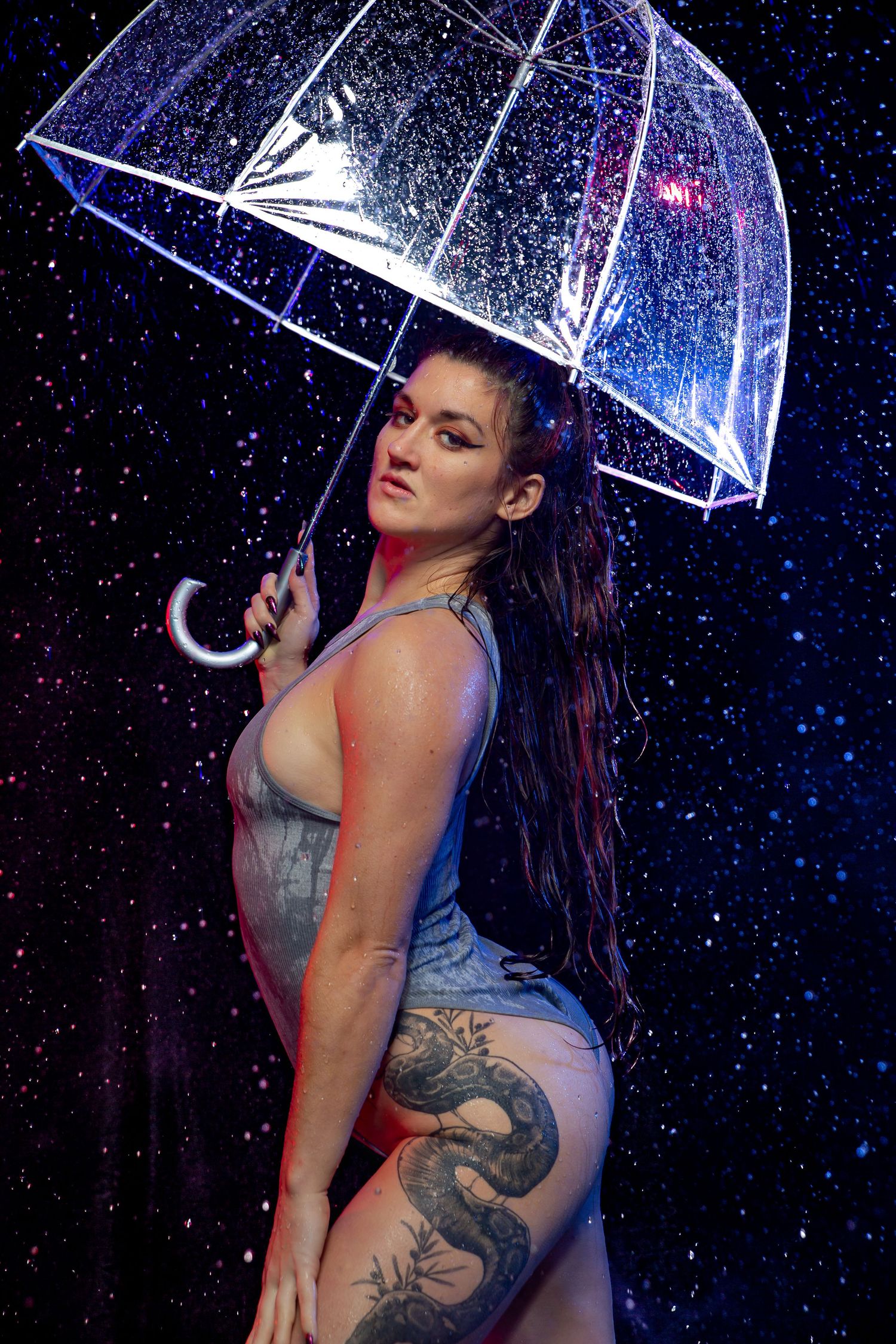 A tattooed woman poses with a clear umbrella in dramatic rain with blue and red studio lighting.