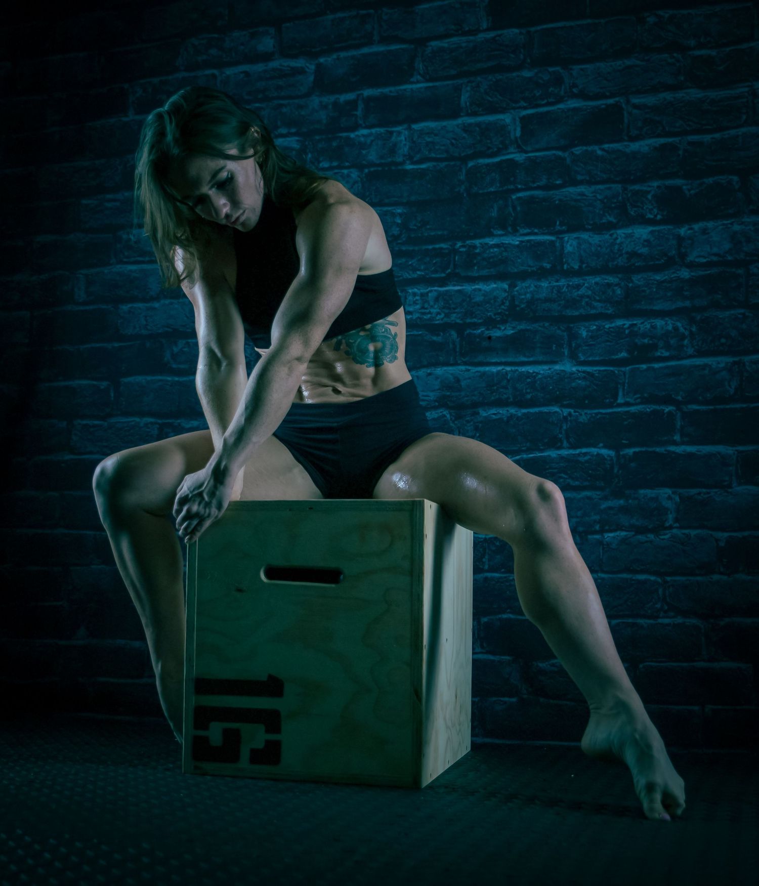Fit woman with tattoos resting on a wooden plyo box in a dark gym with brick wall background.