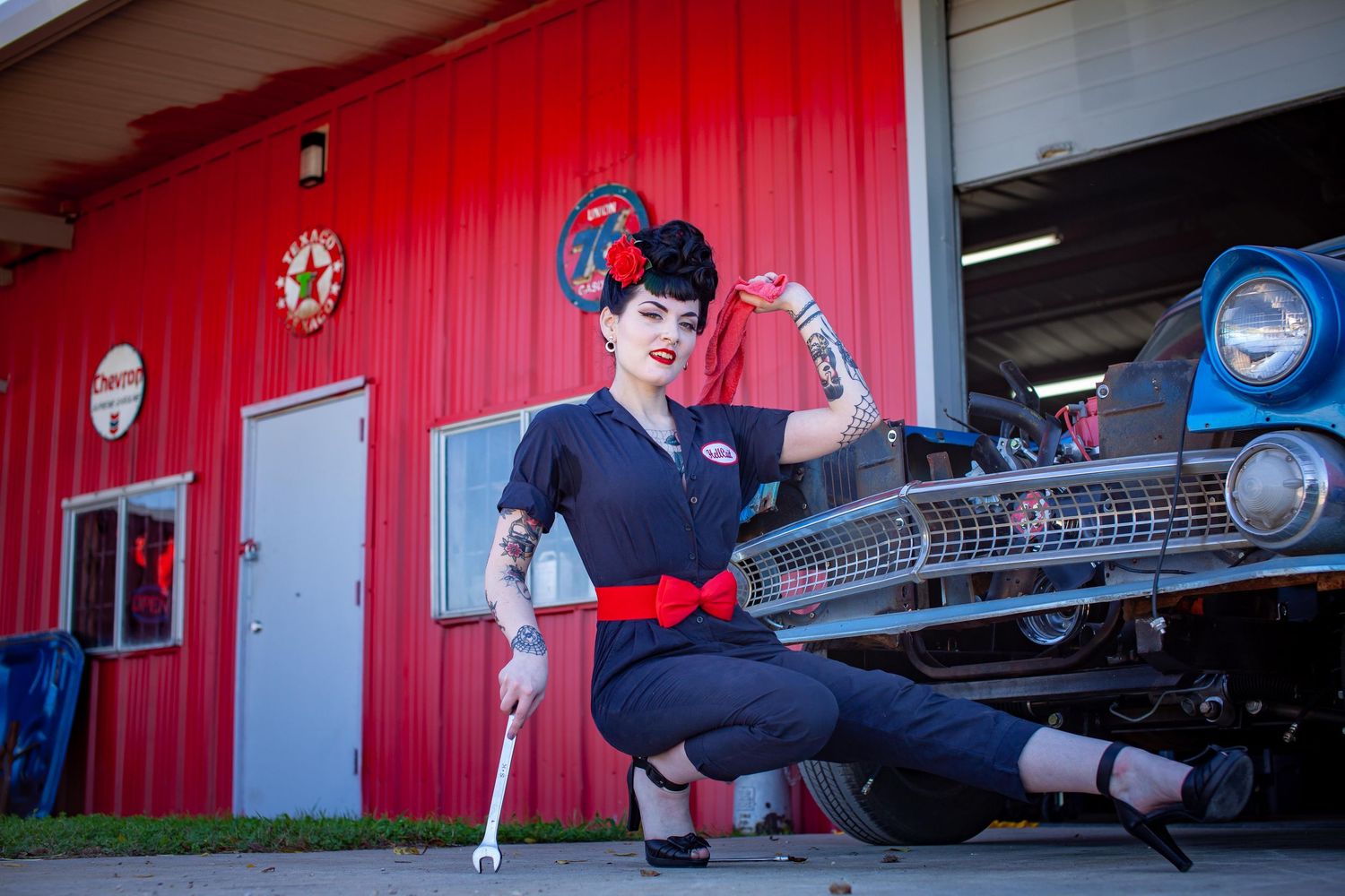 Tattooed pin-up woman in retro mechanic outfit poses with wrench near classic car outside red garage.
