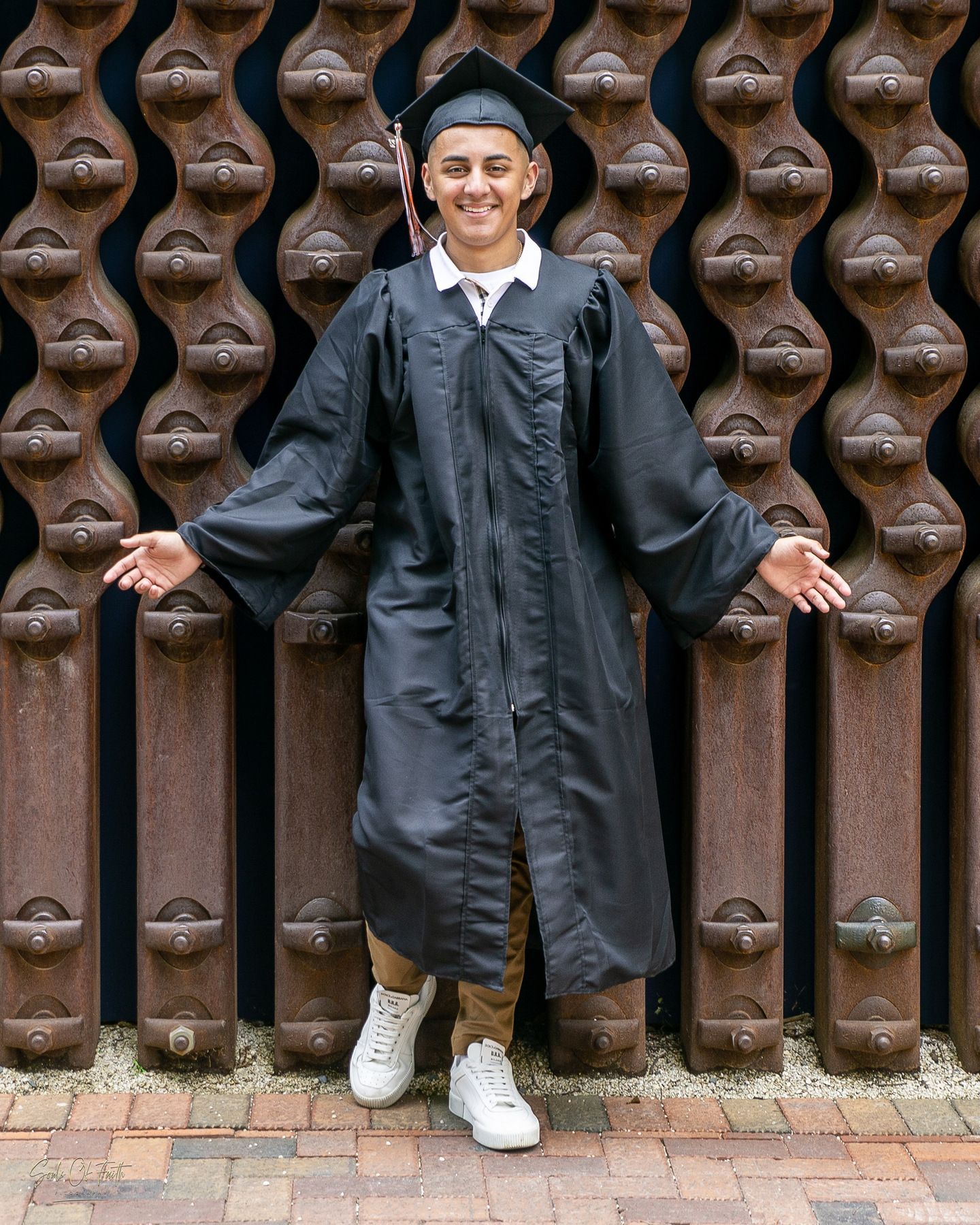 Graduate in black cap and gown smiling with arms outstretched in front of decorative wooden doors.