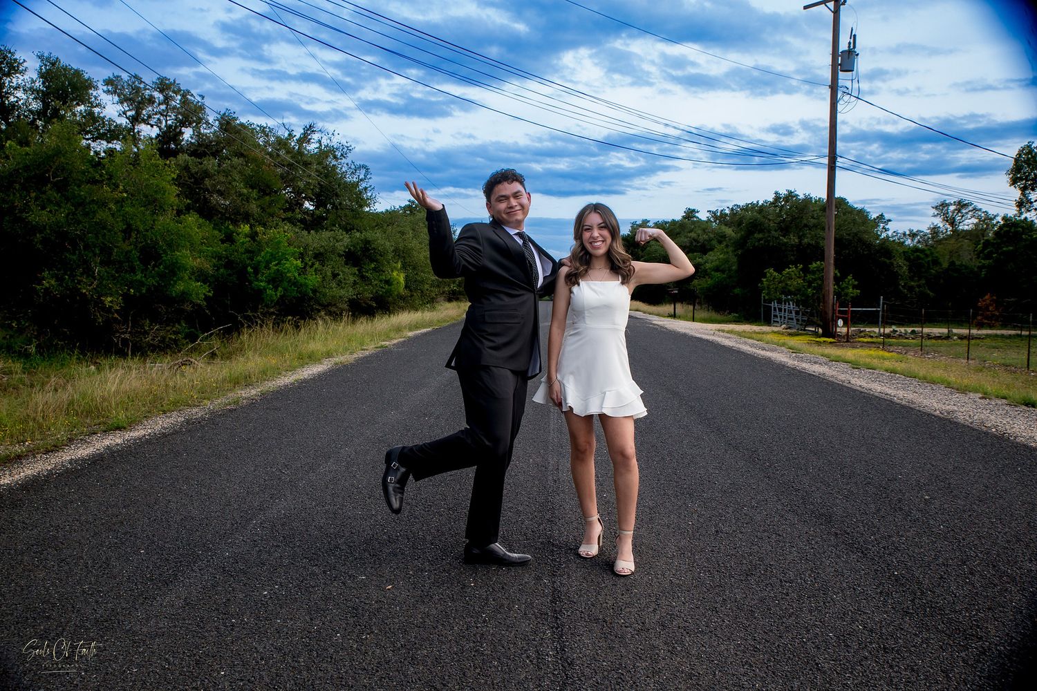 Couple posing playfully on an empty road, man in suit and woman in white dress, with trees and blue sky backdrop.