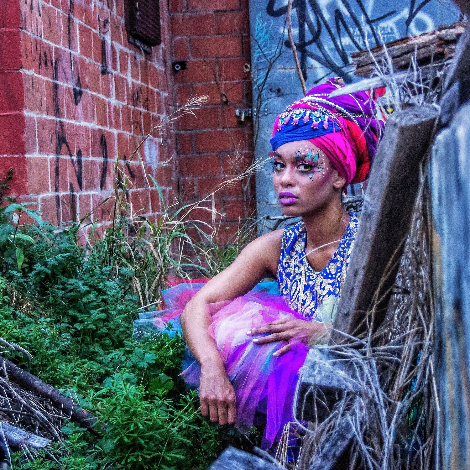 A woman in colorful attire and head wrap poses in an urban alley with graffiti and overgrown plants.