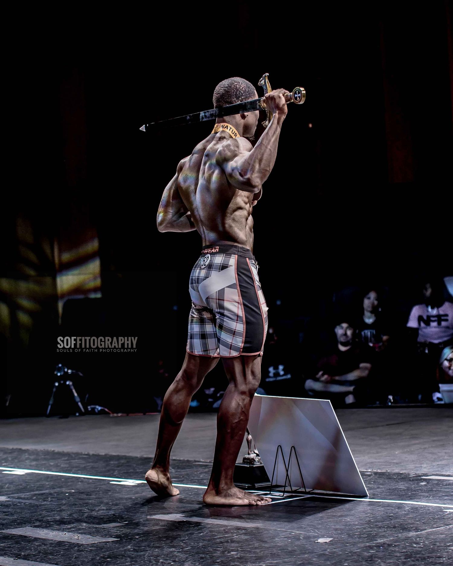 Muscular shirtless male athlete performing on stage with trophy, showcasing back muscles at fitness competition.