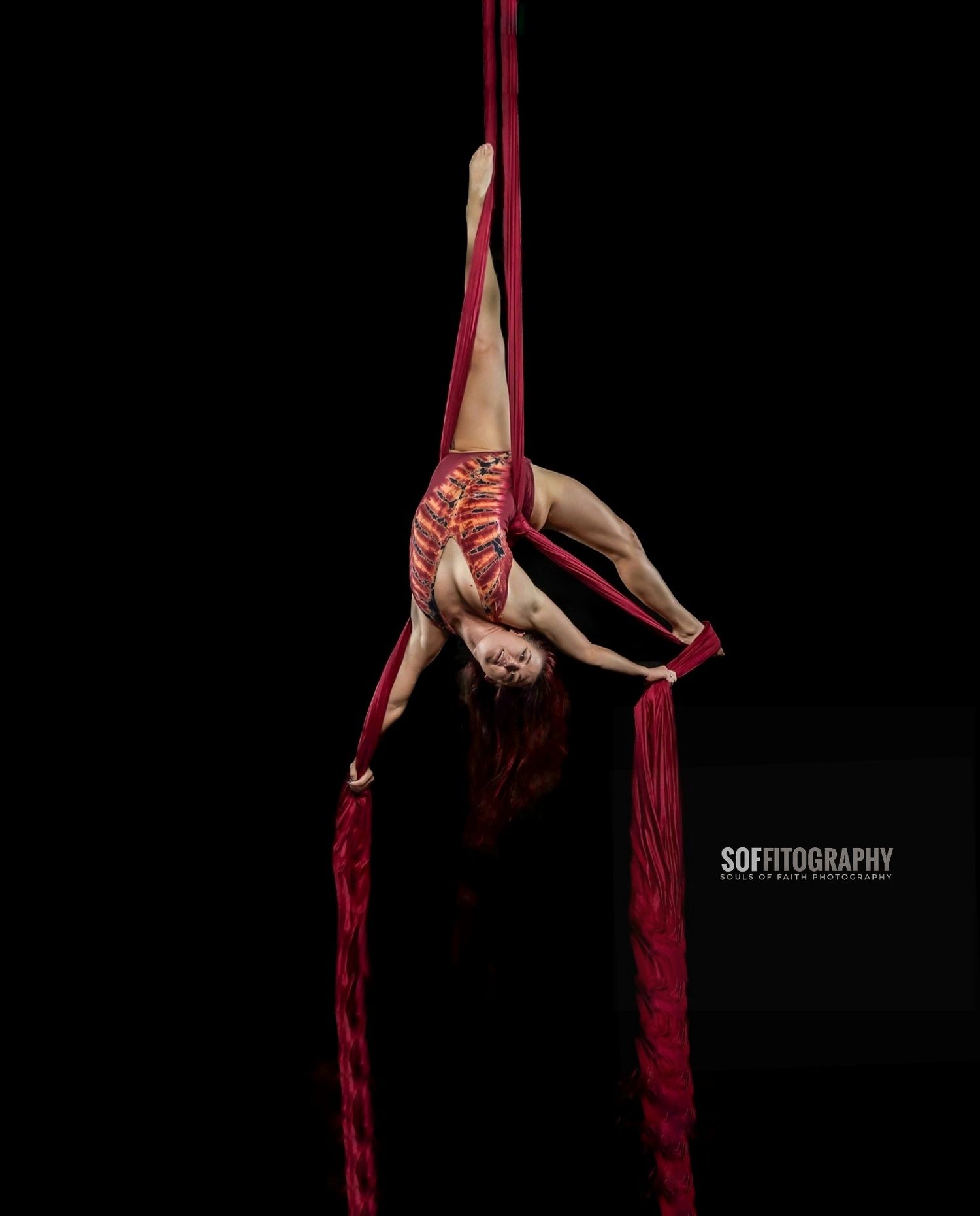 Aerial silk performer in red costume hanging upside down on crimson silks against a dark black background.