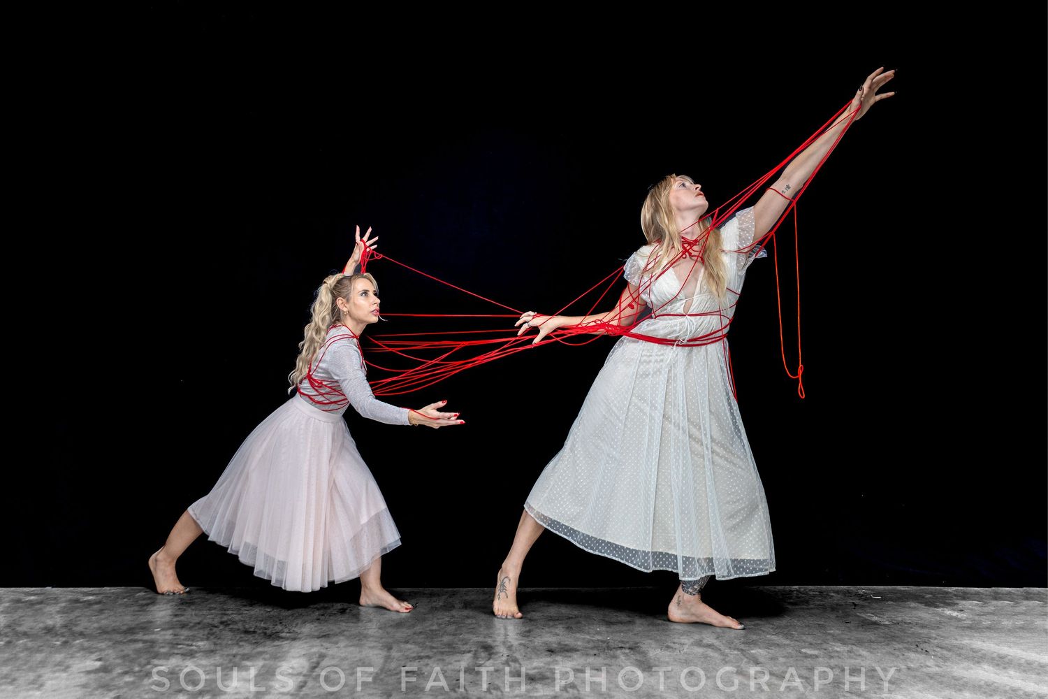 Two dancers in white dresses perform with red ribbons against a dark background in an expressive contemporary dance piece.
