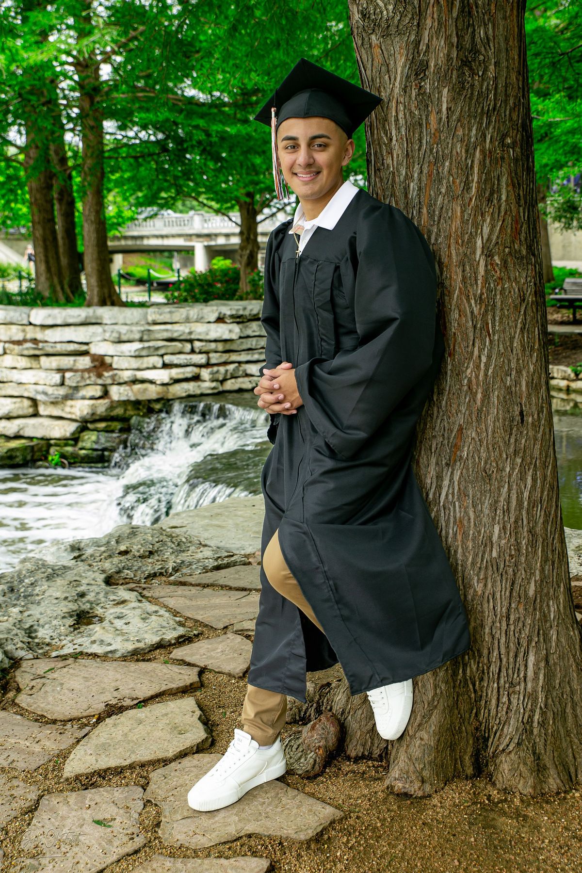 Young man in black graduation cap and gown leaning against a tree, smiling outdoors near a stone waterfall.
