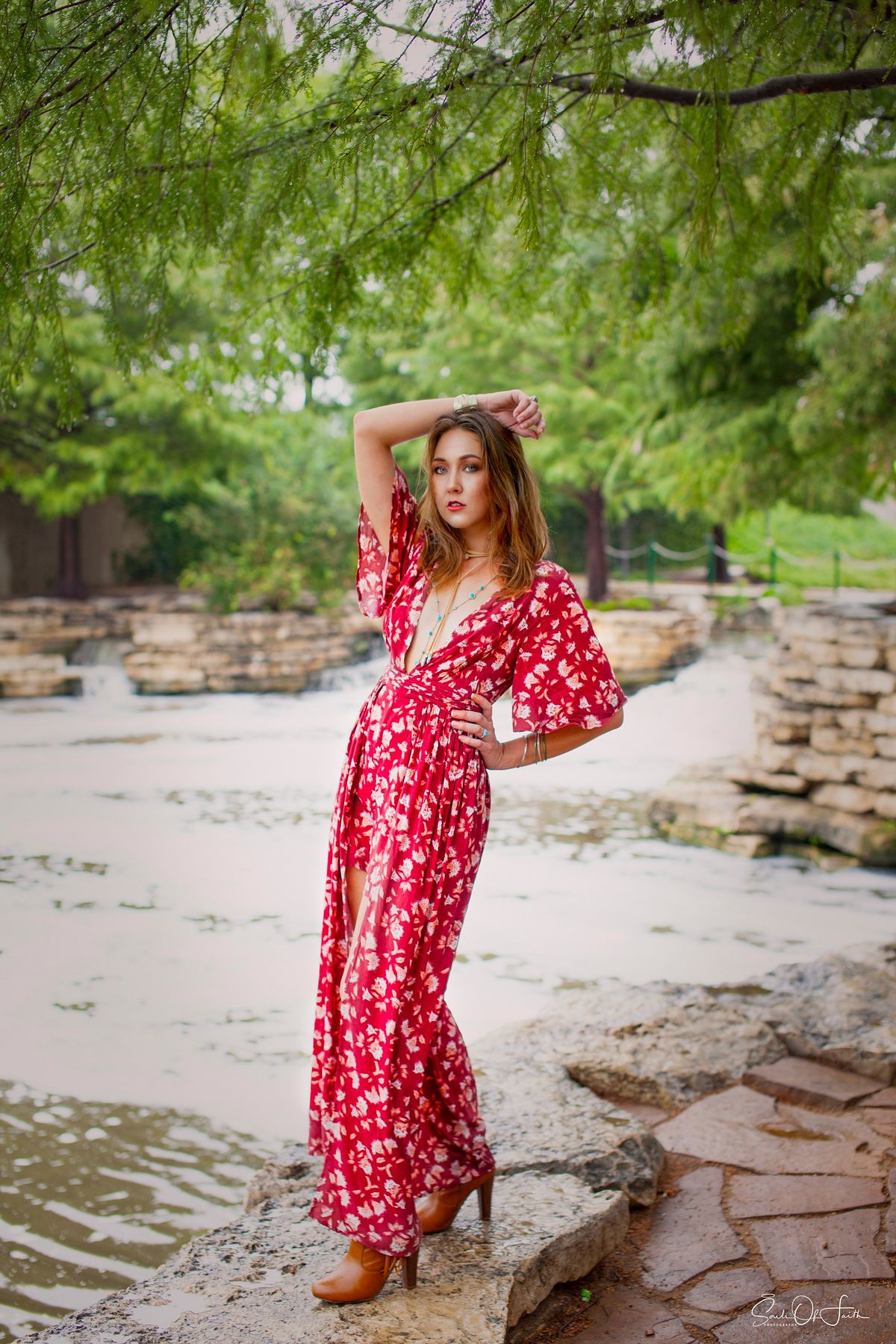 Woman in red floral maxi dress posing on rocks near a pond in a lush green park setting.