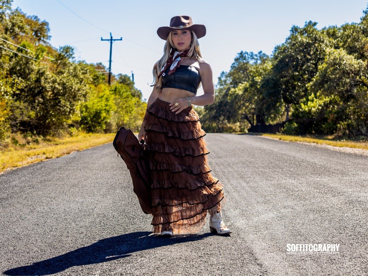 Woman in cowboy hat, brown tiered skirt, crop top, and white boots poses on a rural road surrounded by trees.