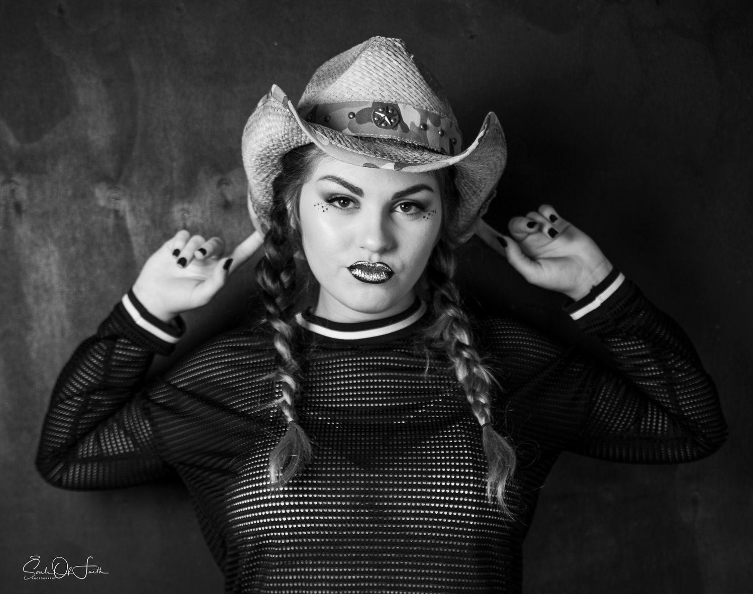 Black and white portrait of a woman in braids and cowboy hat wearing a mesh top, posing confidently.