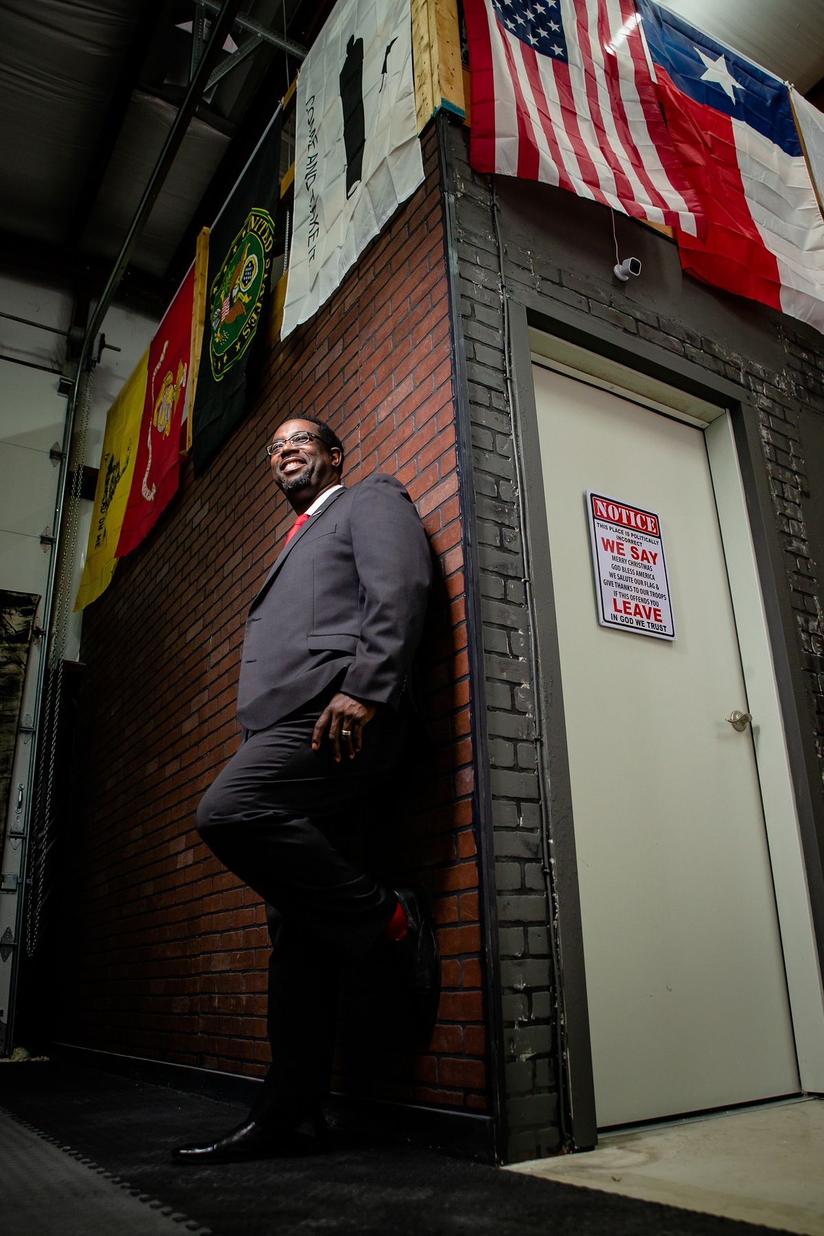 Man in gray suit leaning against brick wall with American and Texas flags hanging above in industrial setting.