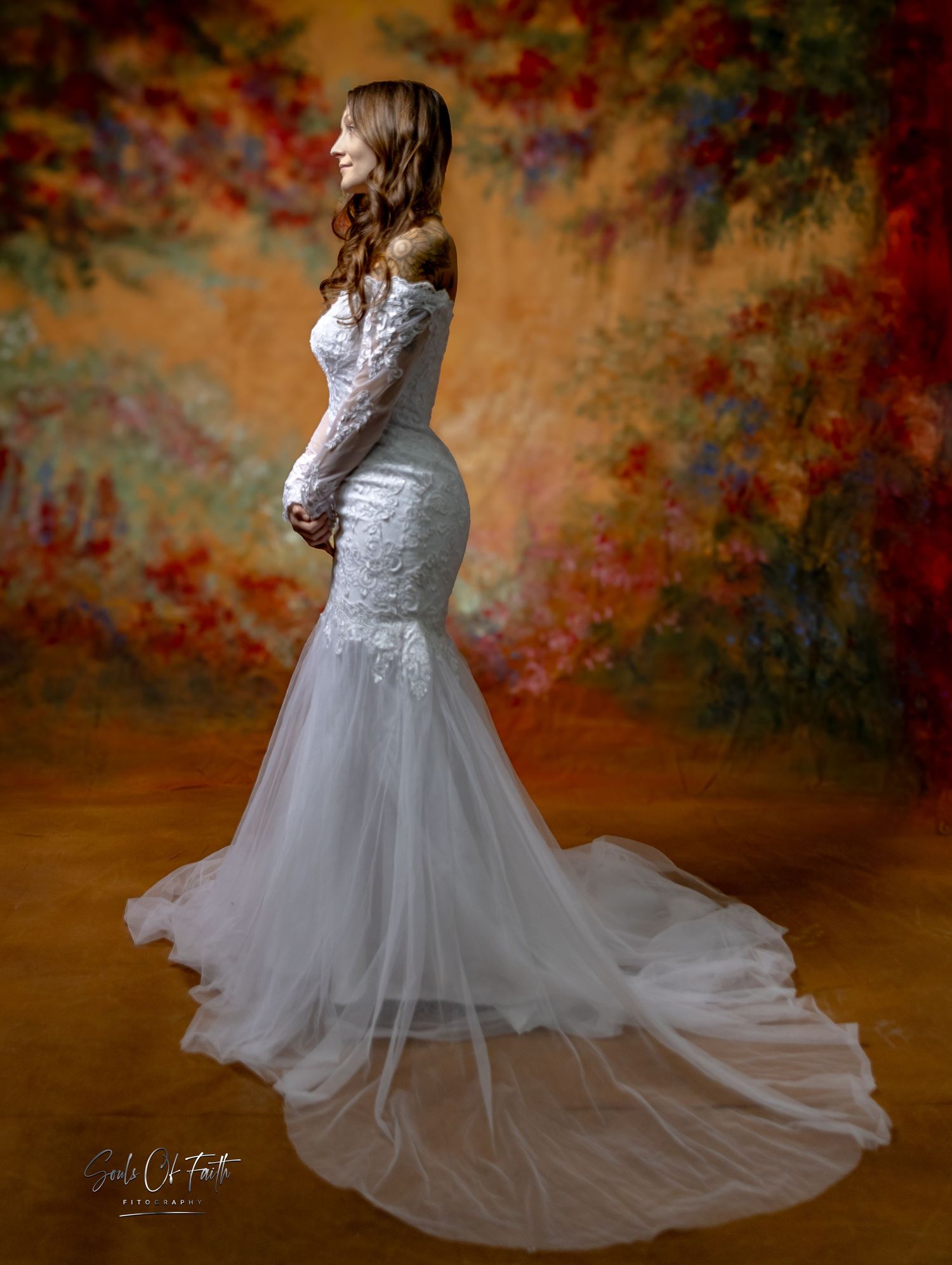 Elegant bride in a white off-shoulder mermaid wedding gown with lace details and tulle train against floral backdrop.
