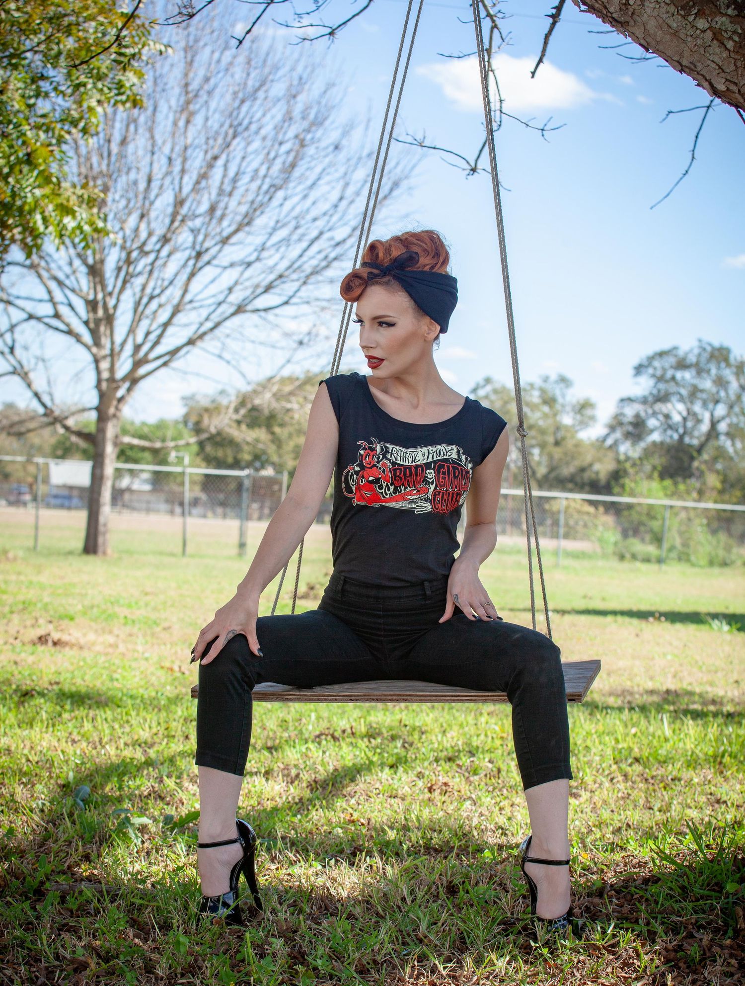 A woman in vintage pin-up style sits on a wooden swing, wearing black pants and graphic tee, with retro hair and heels.