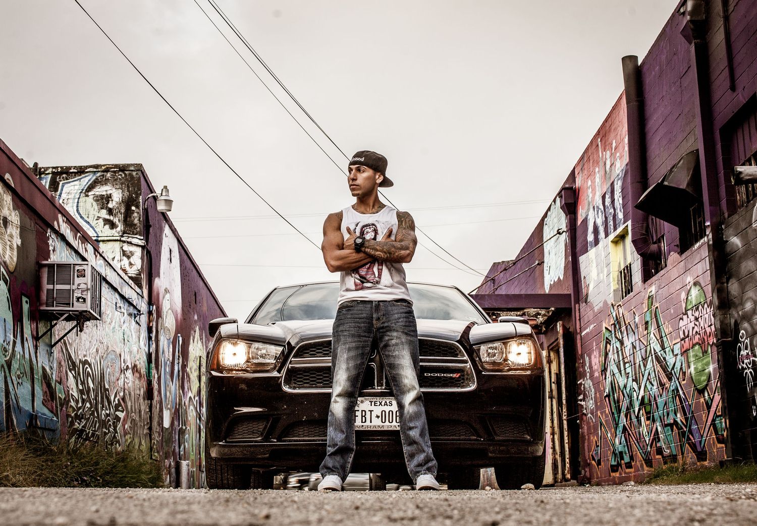 Man in cap and jeans stands arms crossed before a black Dodge Charger in a graffiti-covered urban alley.
