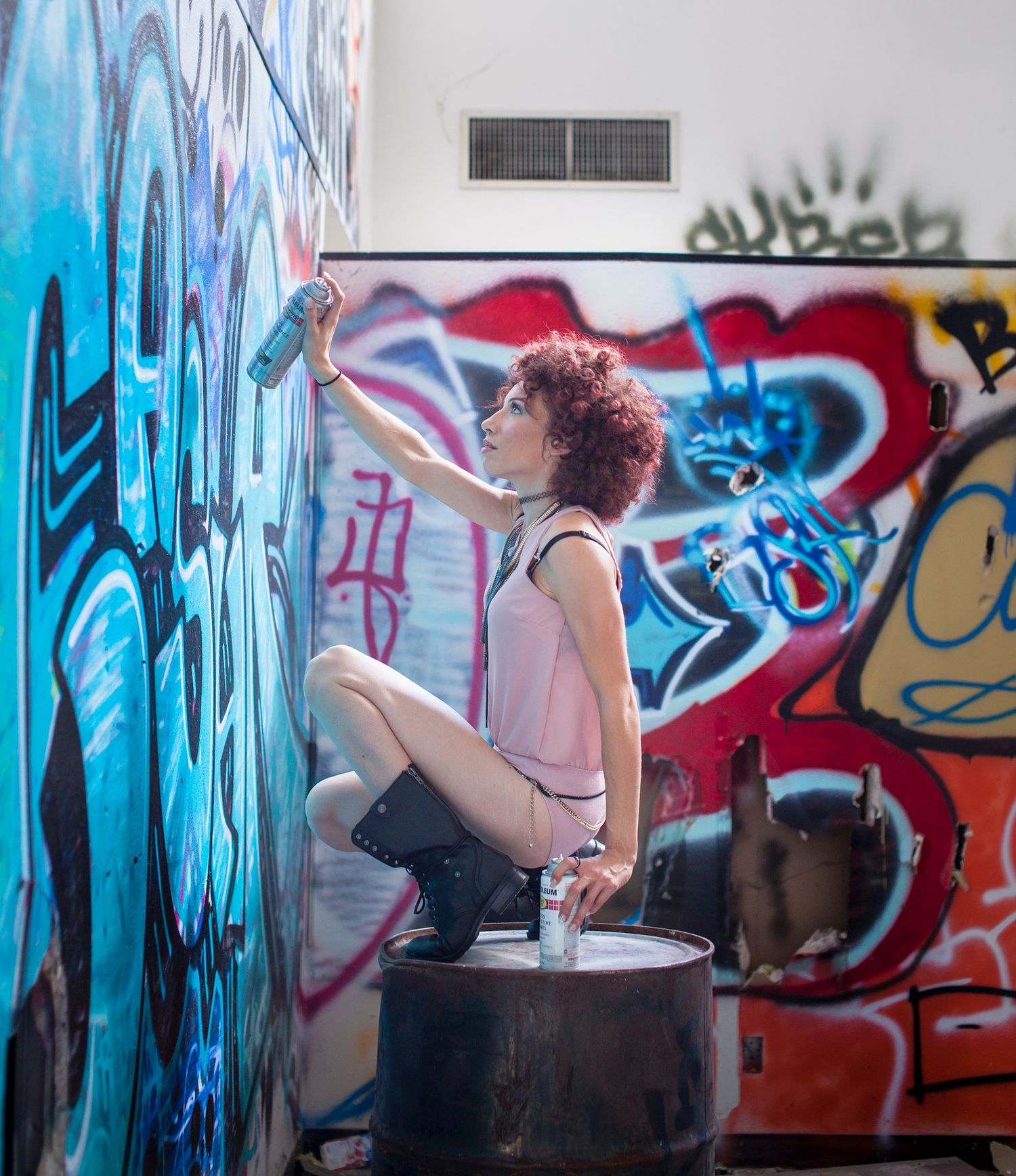 A woman with curly red hair crouches on a bucket, spray painting colorful graffiti art on a wall indoors.
