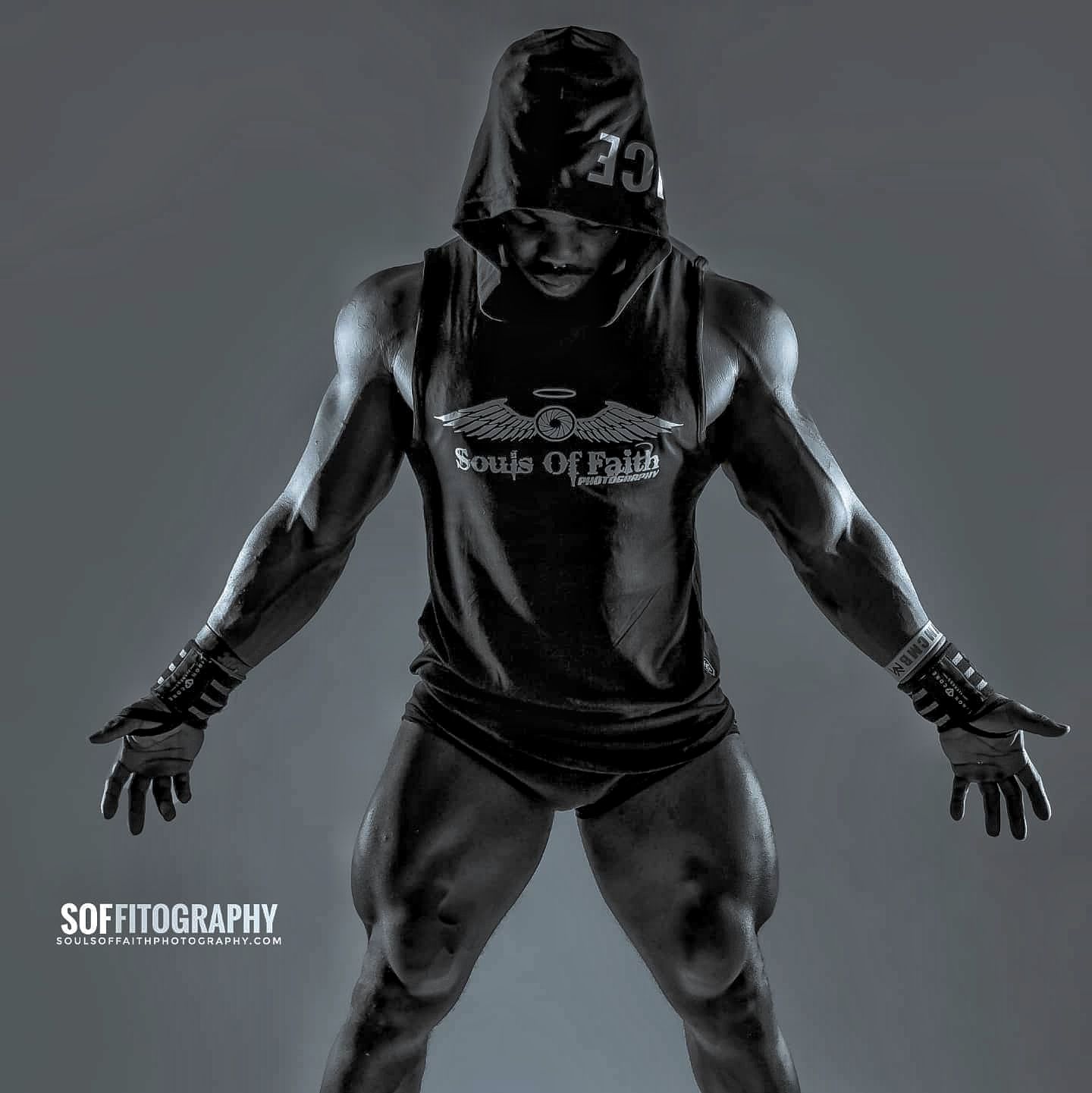 Muscular athlete in hooded tank top and gloves poses dramatically in black and white fitness photo.