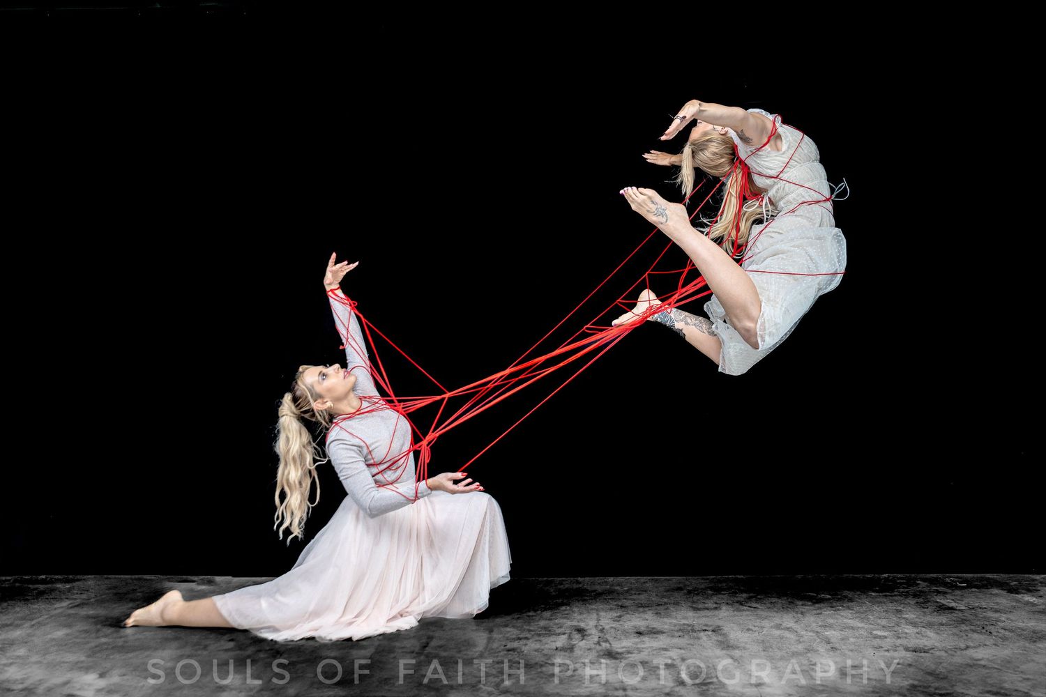 Two dancers perform an acrobatic contemporary dance, connected by red ribbons, one airborne and one on the floor.