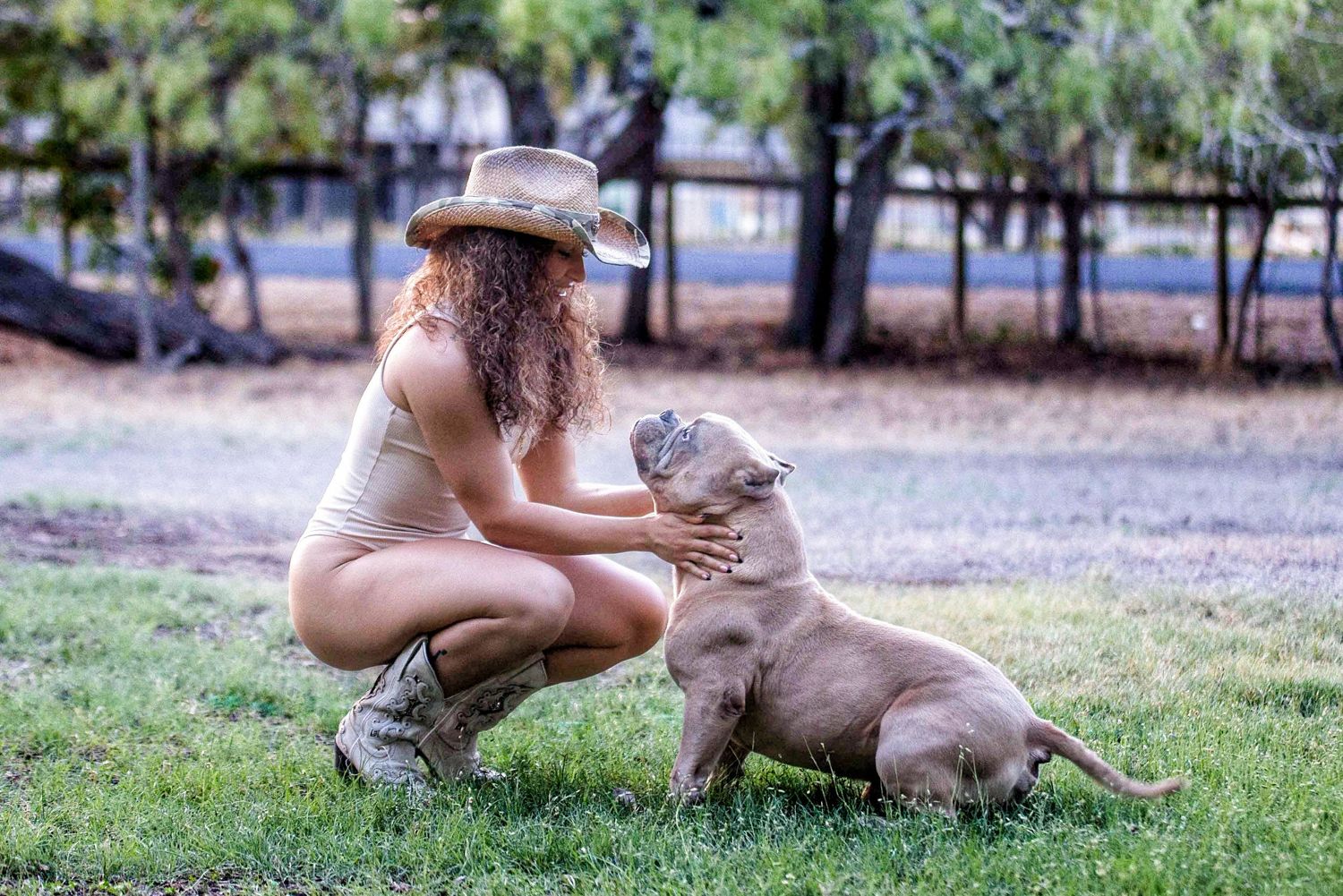 Woman in cowboy hat crouching on grass, affectionately petting a young pit bull puppy outdoors near a fence.